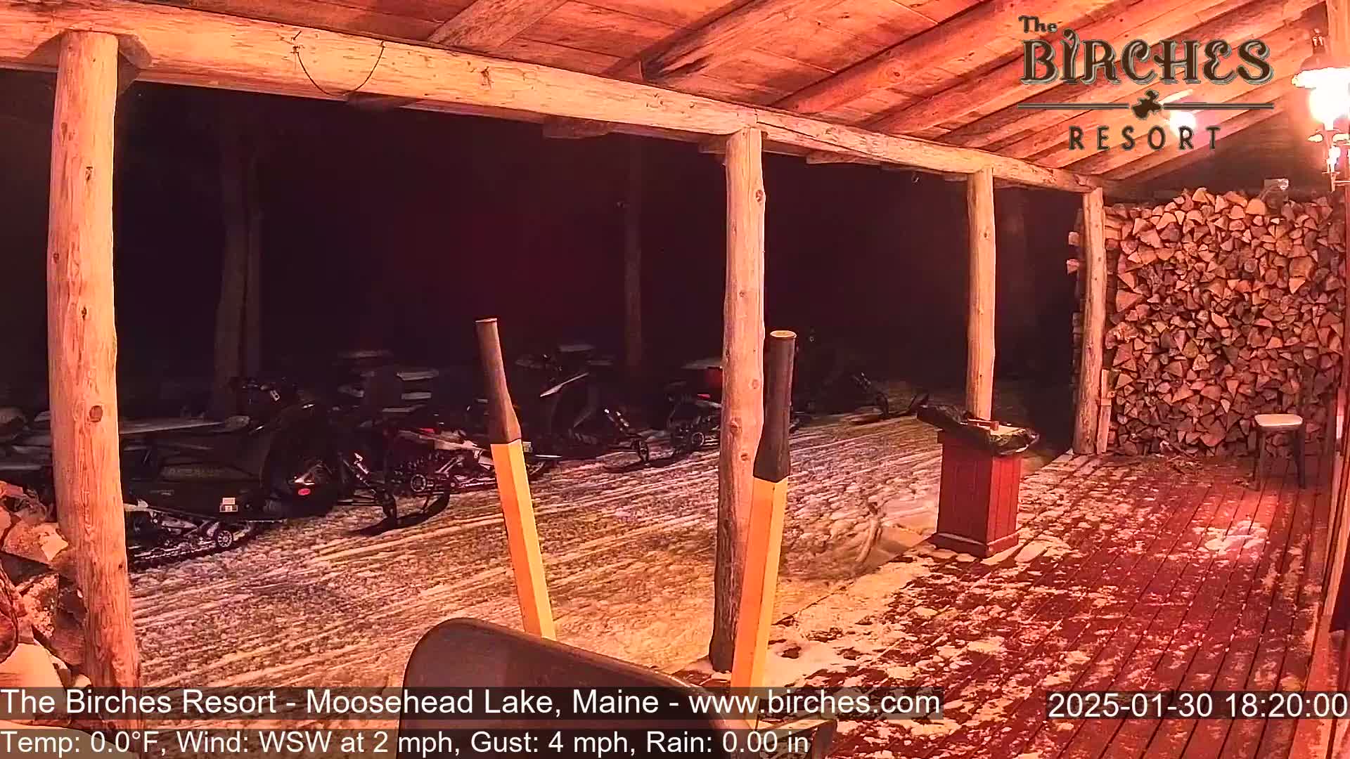 A snow-covered wooden porch at night, with snowmobiles parked outside and a woodpile visible to the right.