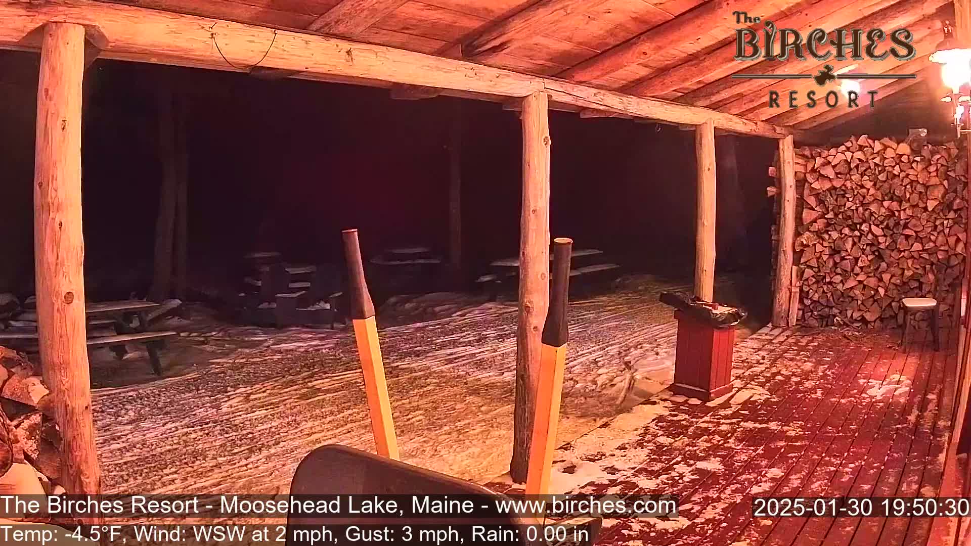 A snow-covered outdoor patio with picnic tables is viewed from under a wooden covered porch at night, with a woodpile visible to the right.
