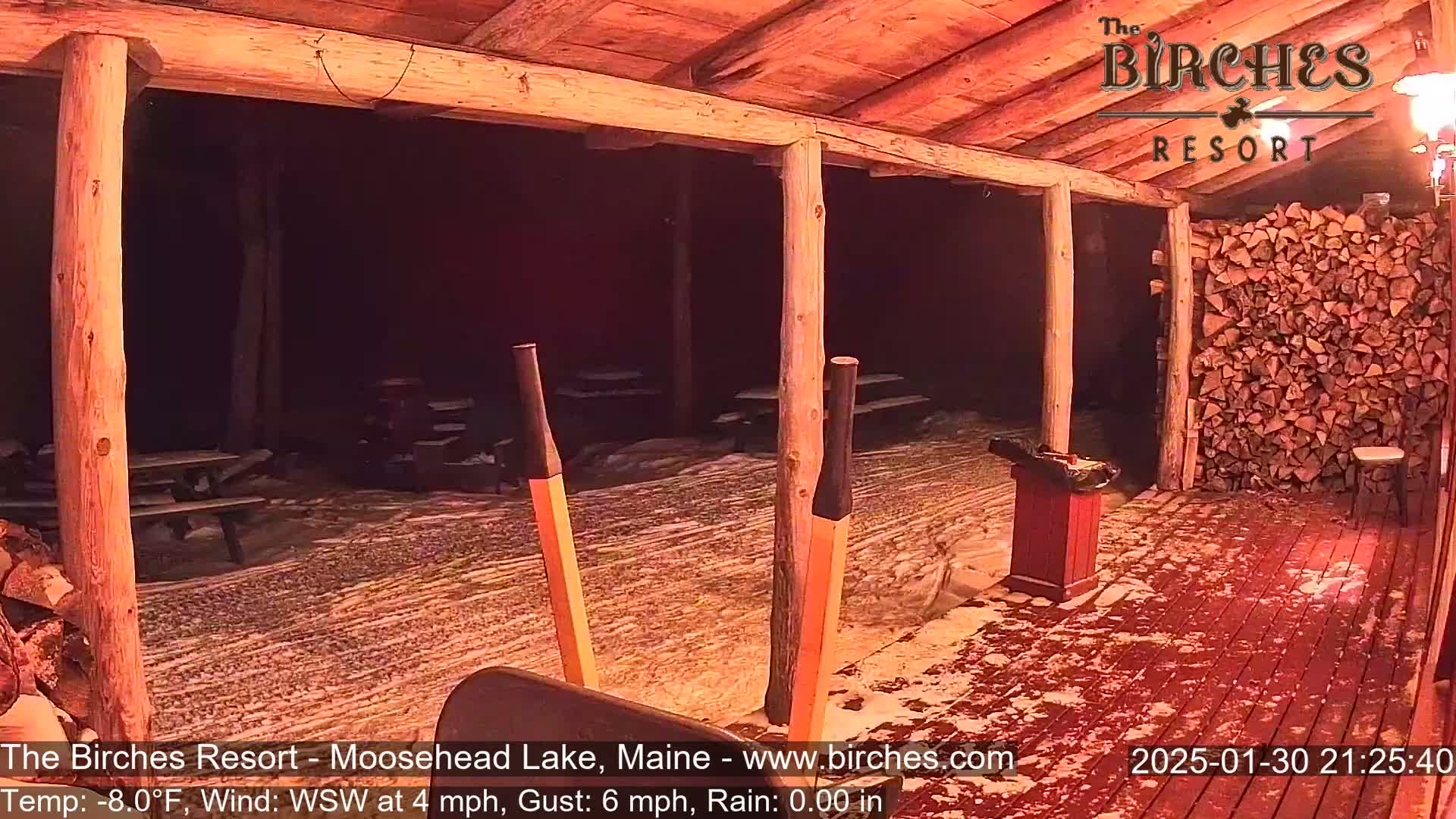 A snow-covered outdoor patio with picnic tables and a woodpile is viewed from inside a rustic wooden structure at night.