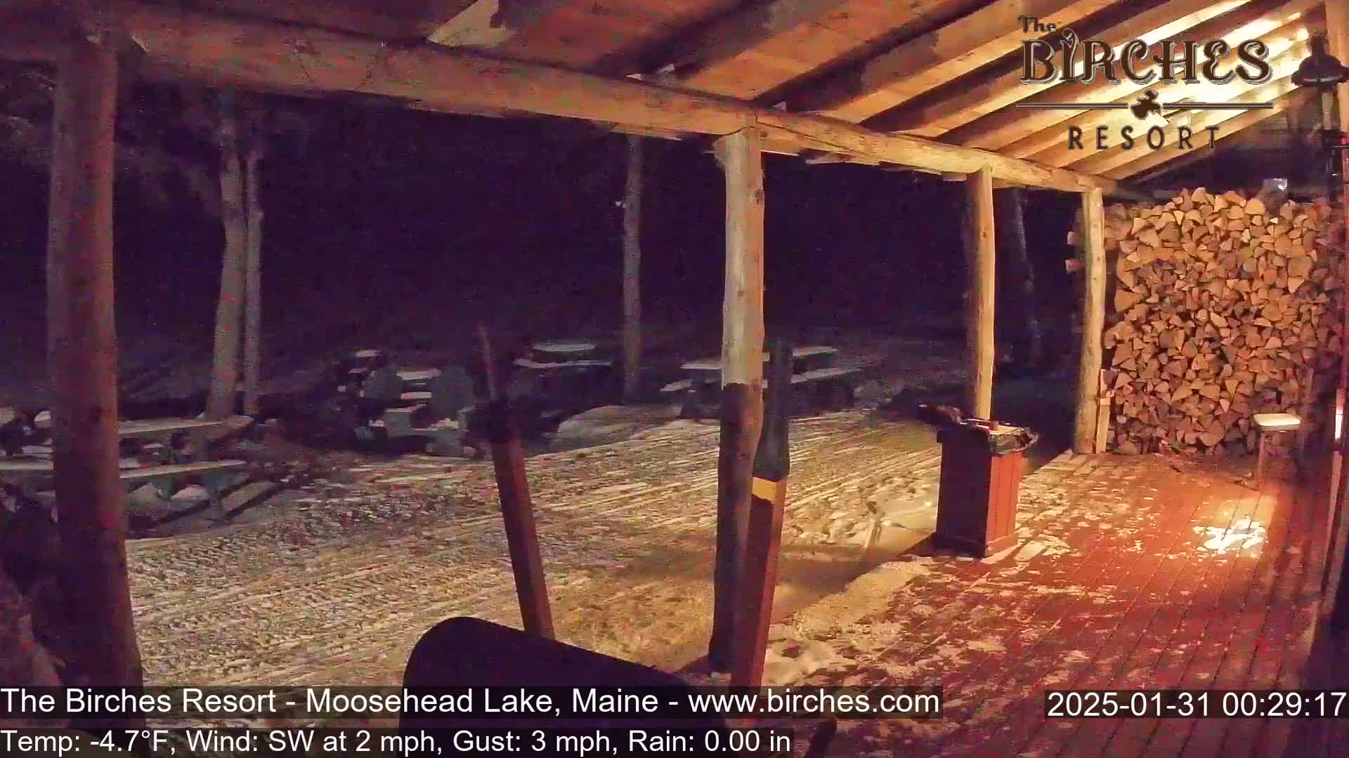 A snow-covered outdoor patio area with picnic tables under a wooden structure at night, illuminated by a light near a stack of firewood.