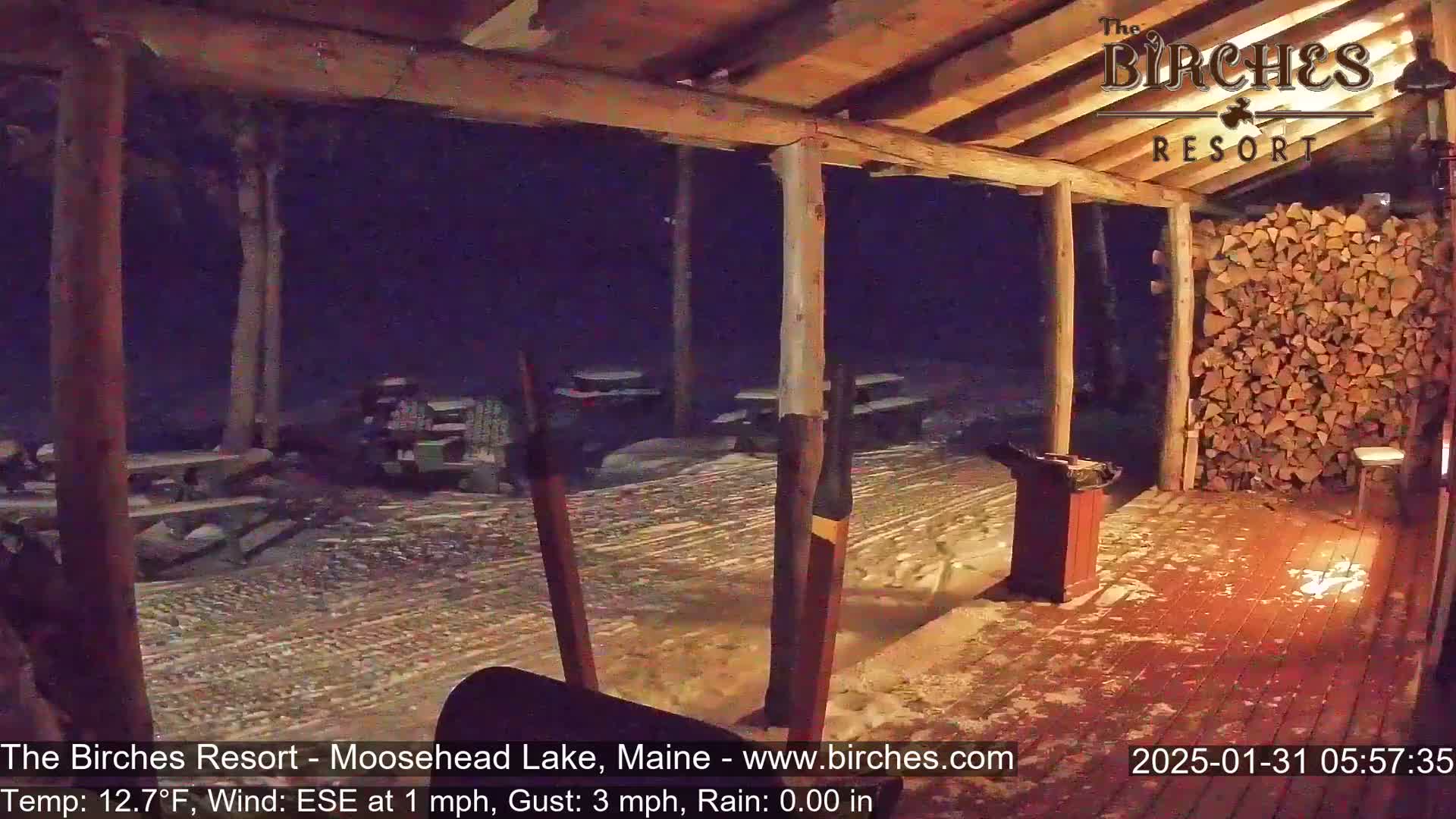 A snow-covered porch with picnic tables and a woodpile is viewed from inside a building at night under rainy conditions.