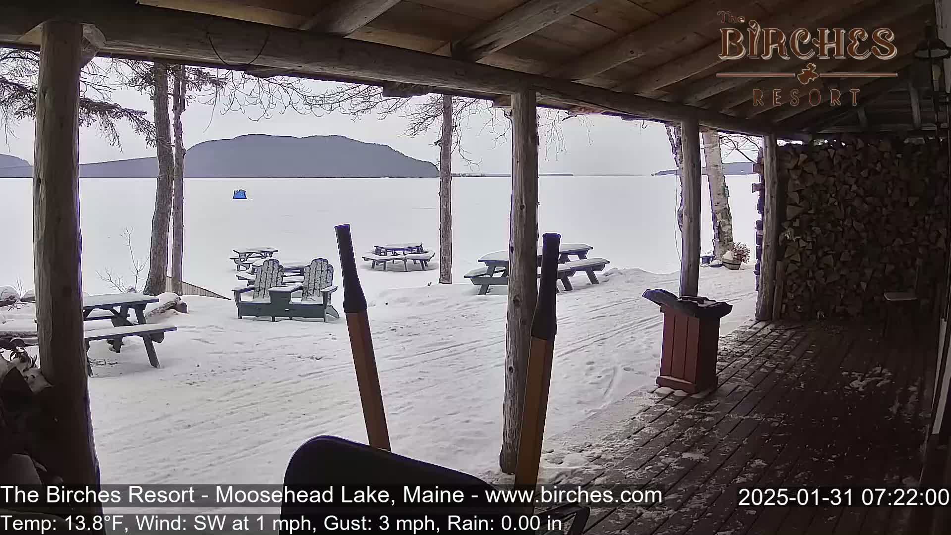 A snow-covered patio with picnic tables and Adirondack chairs overlooks a frozen lake and distant mountains under an overcast sky.