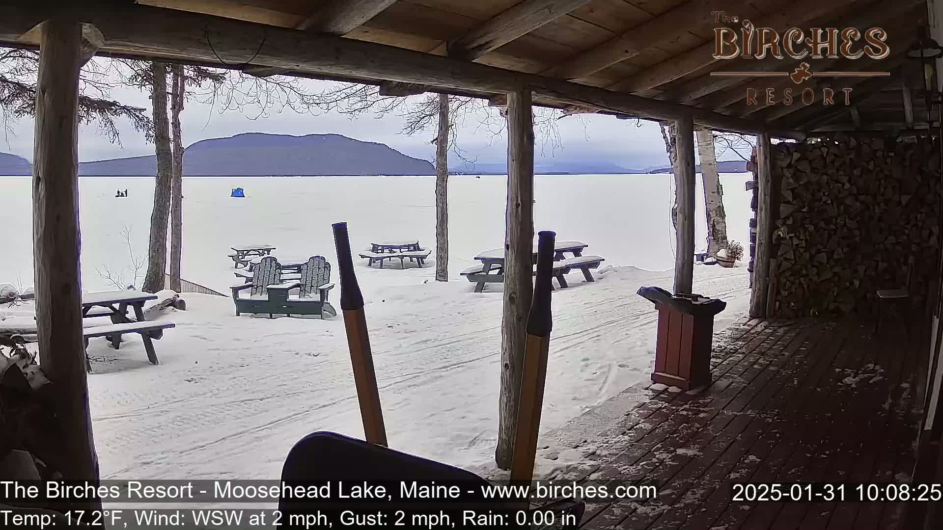 A snow-covered porch overlooks a frozen lake with distant mountains under an overcast sky.