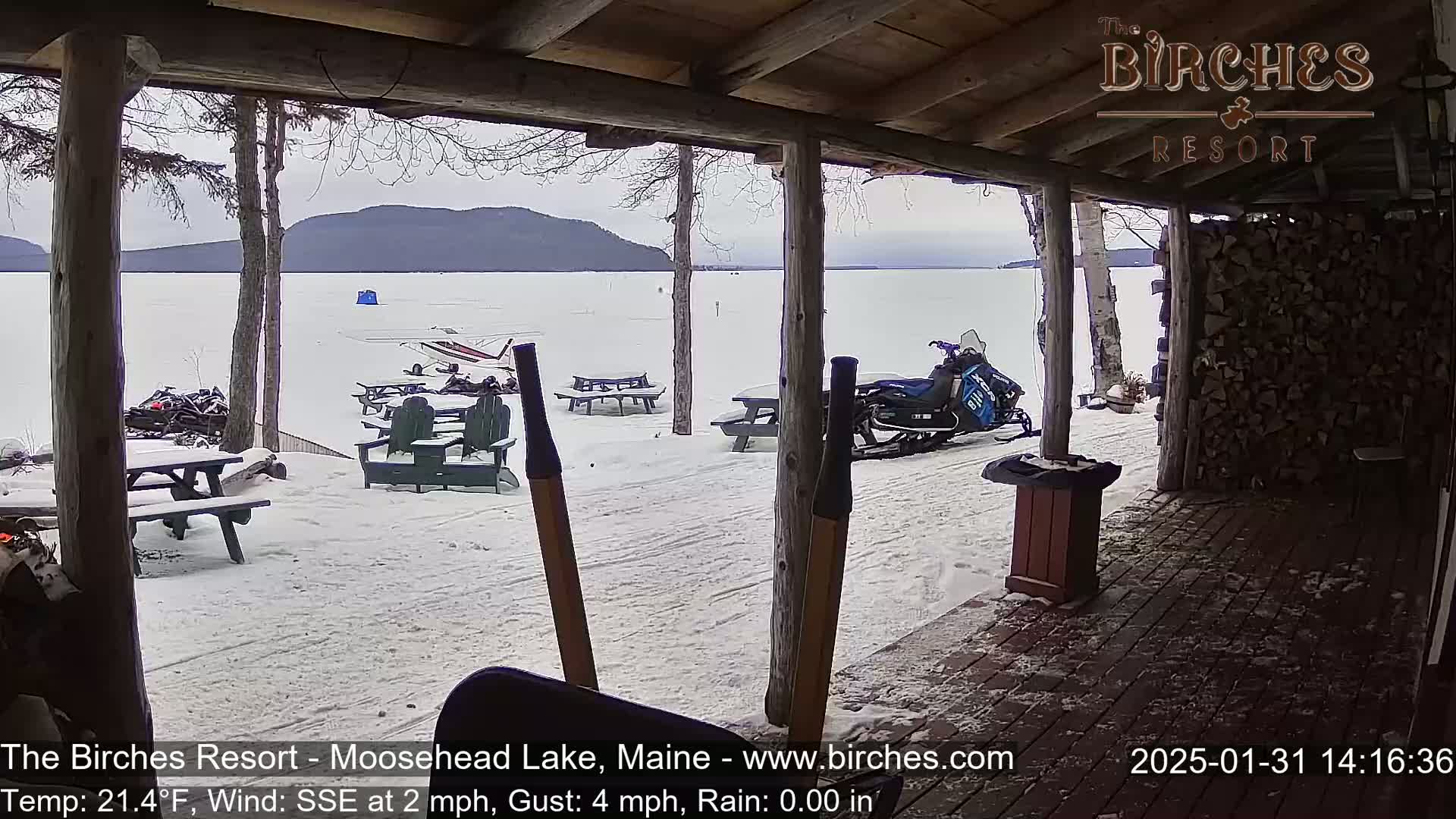 A snow-covered lakeside view is seen from a covered porch, with picnic tables, chairs, and a snowmobile present in the lightly falling rain.