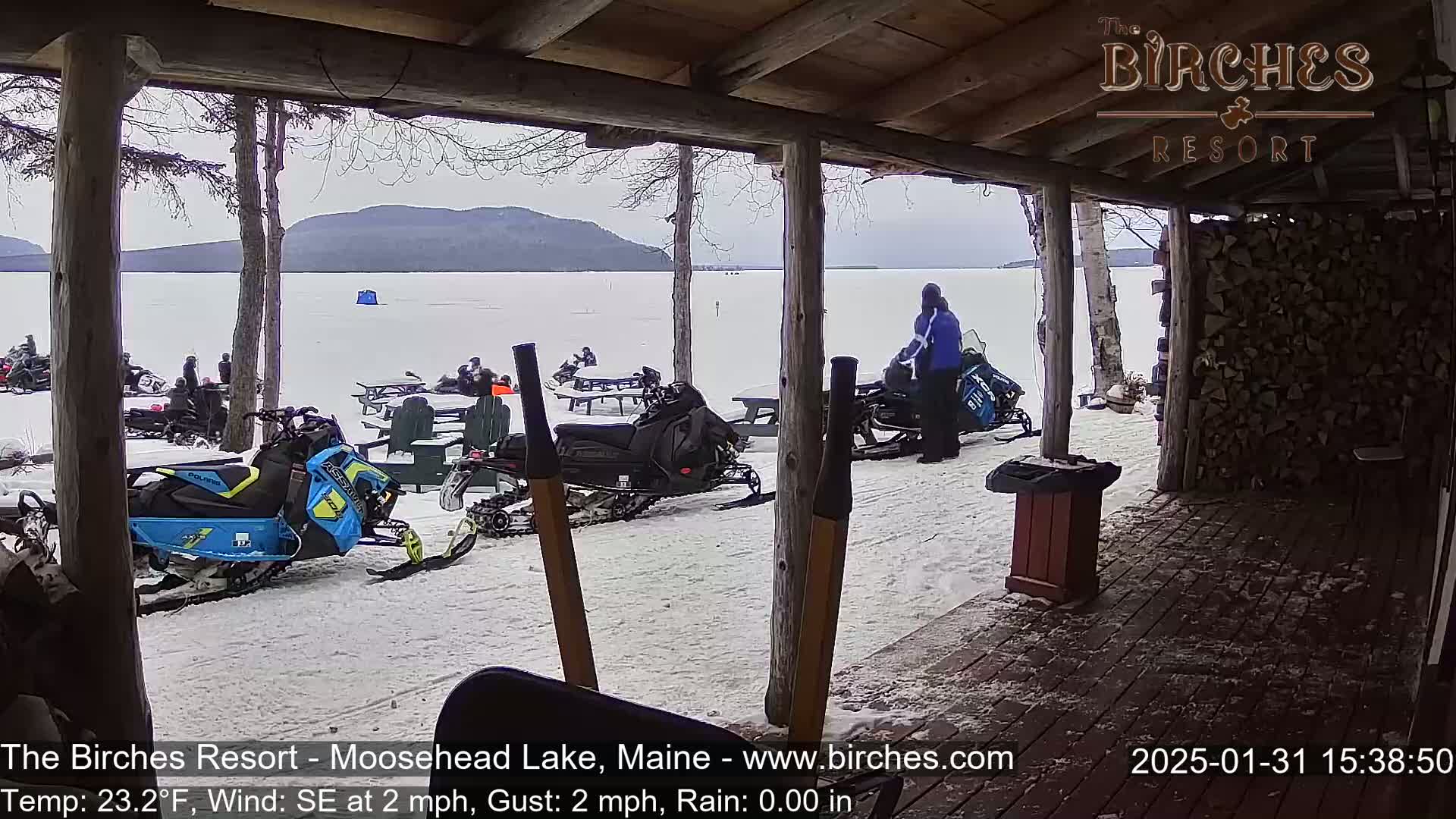From a snow-covered porch overlooking a frozen lake with mountains in the distance under an overcast sky, several snowmobiles are parked near people sitting at picnic tables.