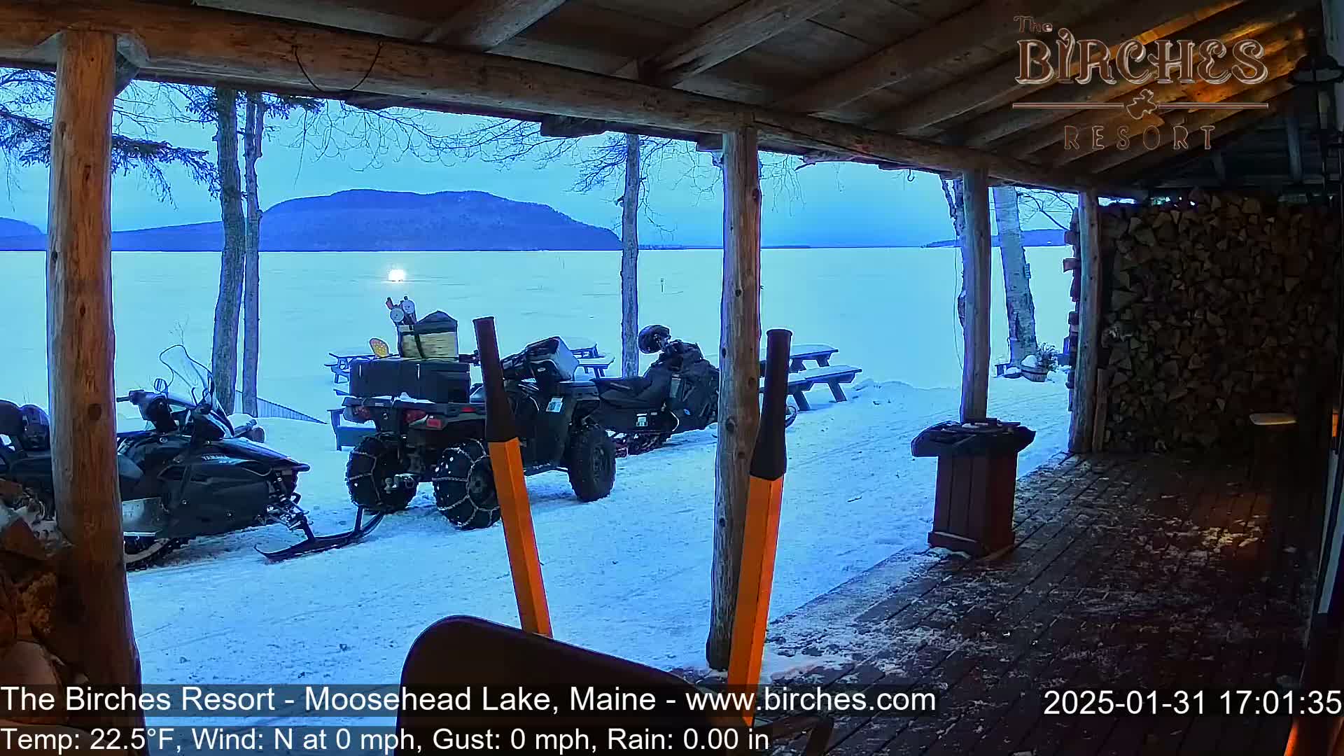 From a wooden porch overlooking a snow-covered lake and distant mountains under a twilight sky, two snowmobiles and an ATV are parked.