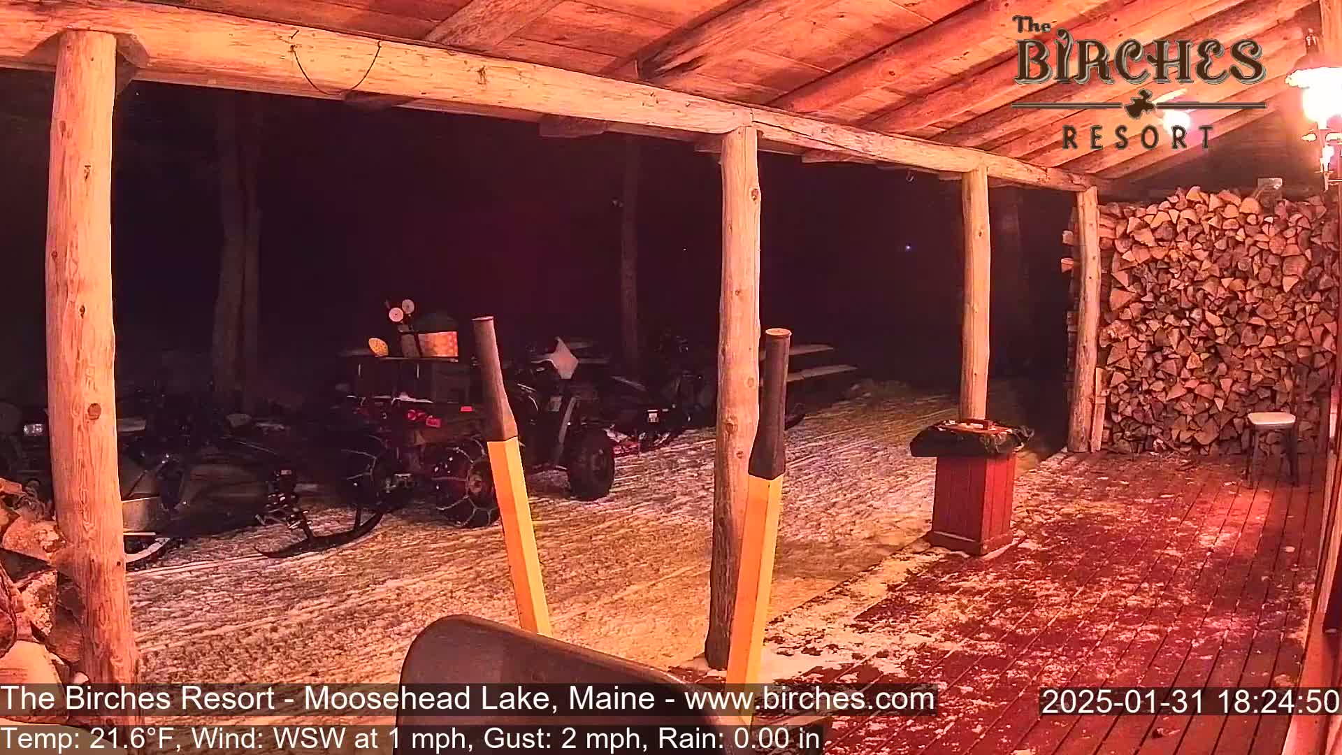 A snow-covered outdoor area with several snowmobiles parked under a wooden porch at night, illuminated by a warm light, with a stack of firewood visible to the right.