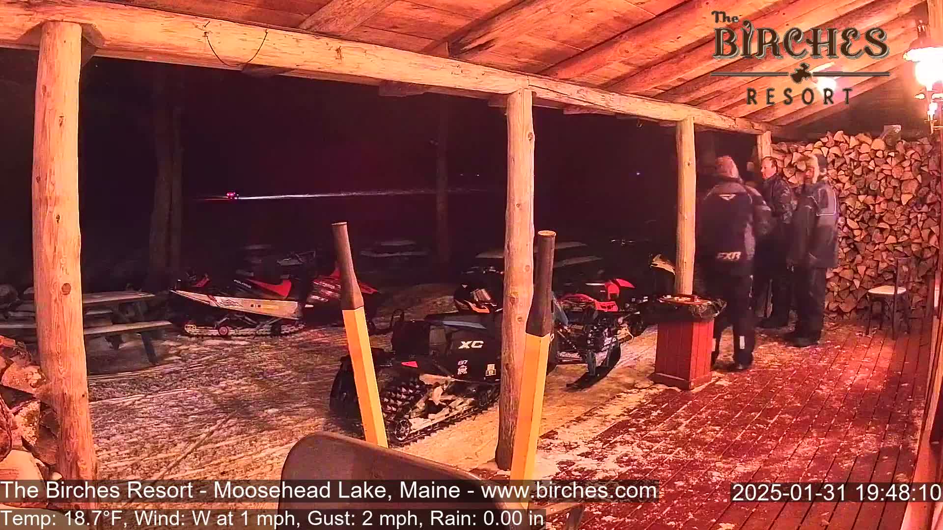 Under a covered porch at night in light rain, several snowmobiles are parked near three people standing by a woodpile.