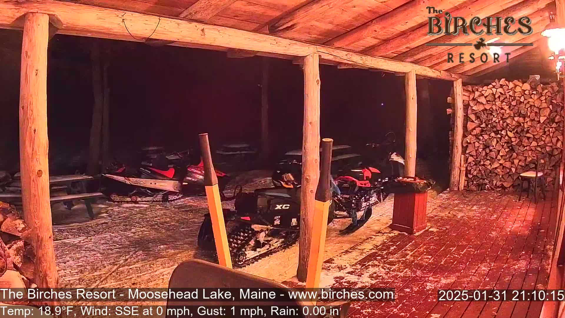 Under a covered porch at night in the snow, several snowmobiles are parked near a woodpile and picnic tables.