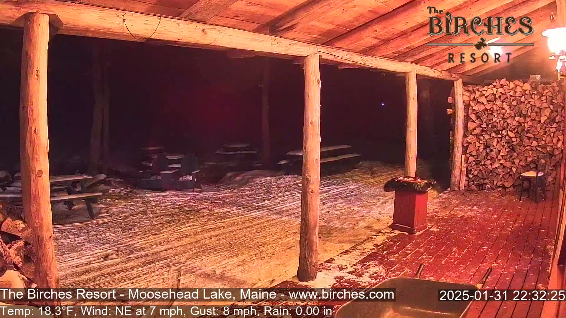 A snow-covered outdoor area with picnic tables is viewed from a wooden porch at night, under a northeast wind.
