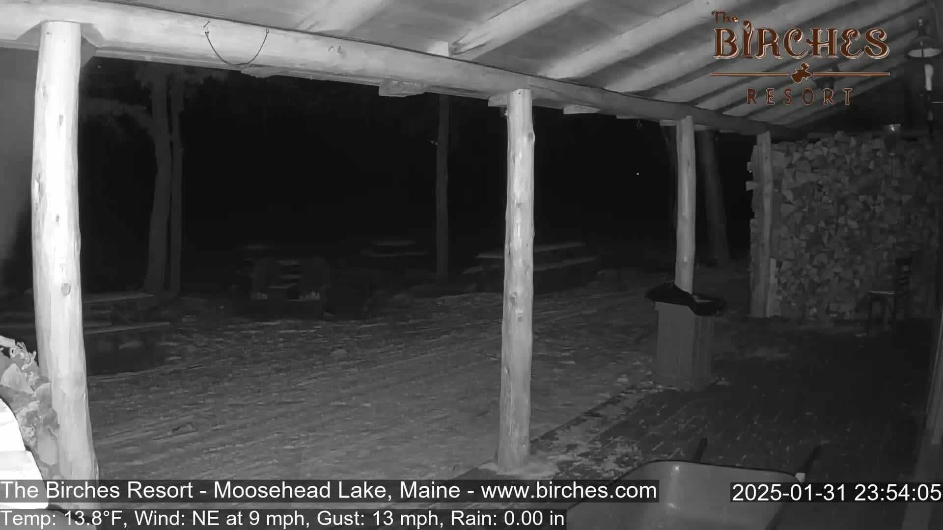 A snow-covered outdoor patio area is visible at night under a wooden structure, with a woodpile nearby, under a northeast wind.
