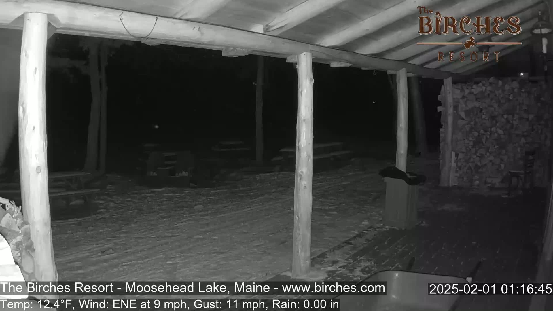 A snow-covered outdoor patio area at night, under a covered porch with wooden posts, is lit by a security camera.