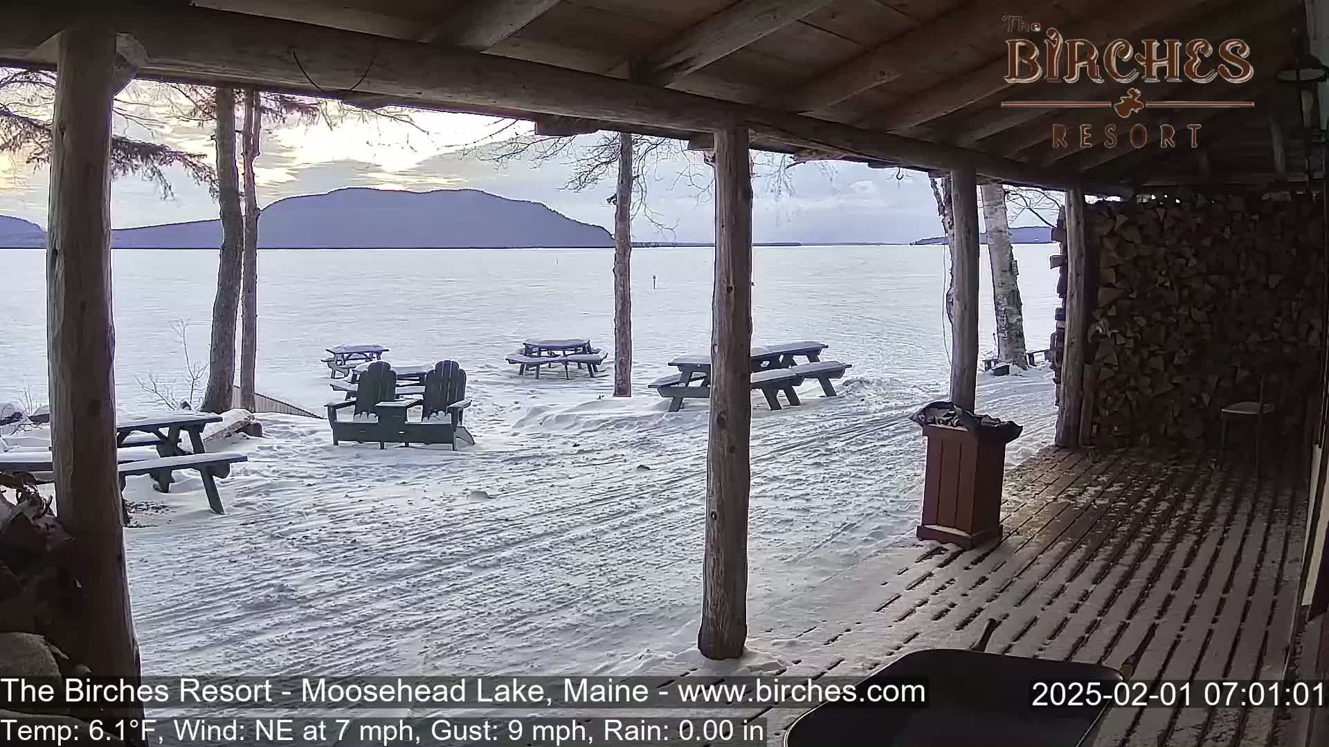 A snow-covered lakeside patio with picnic tables and chairs is seen from inside a wooden structure overlooking a calm lake and distant mountains under a cloudy sky.