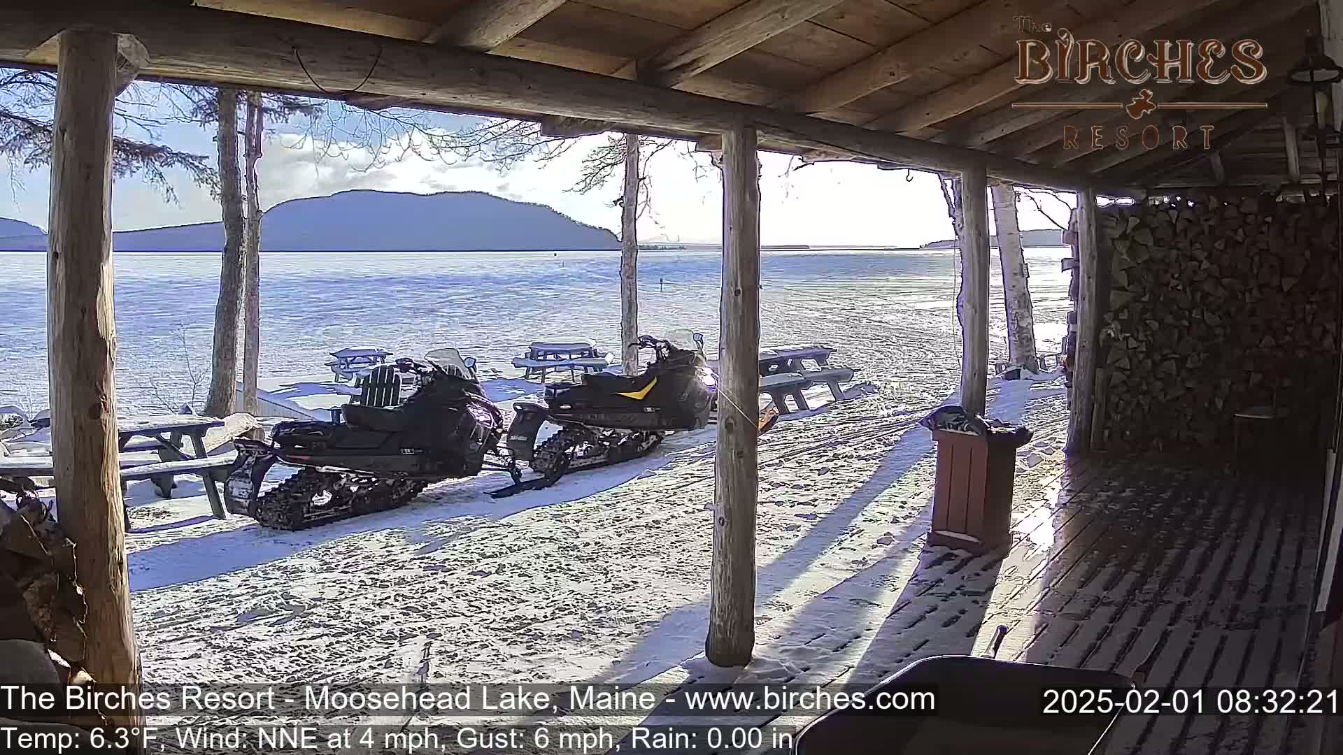 From a snow-covered porch overlooking a calm lake and distant mountains, two snowmobiles are parked near picnic tables under a partly sunny sky.