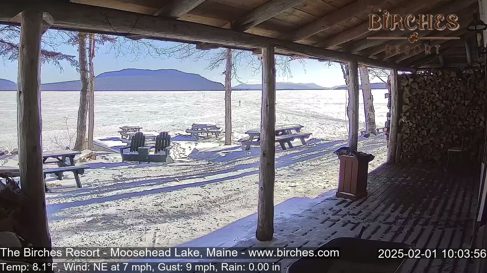 A view from a snow-covered porch shows a partly frozen lake, distant mountains, and several picnic tables under a sunny sky.