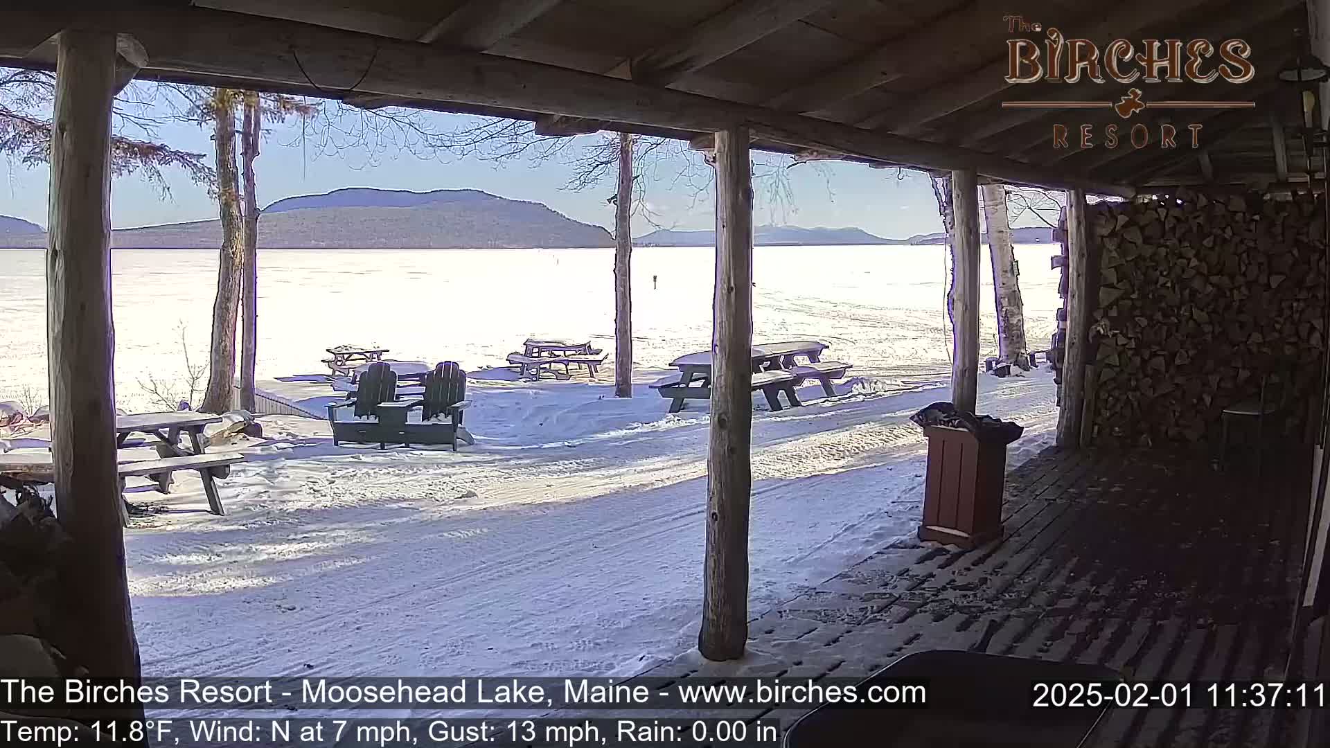A snow-covered, frozen lake is visible from a wooden porch with picnic tables and Adirondack chairs, under a sunny sky.