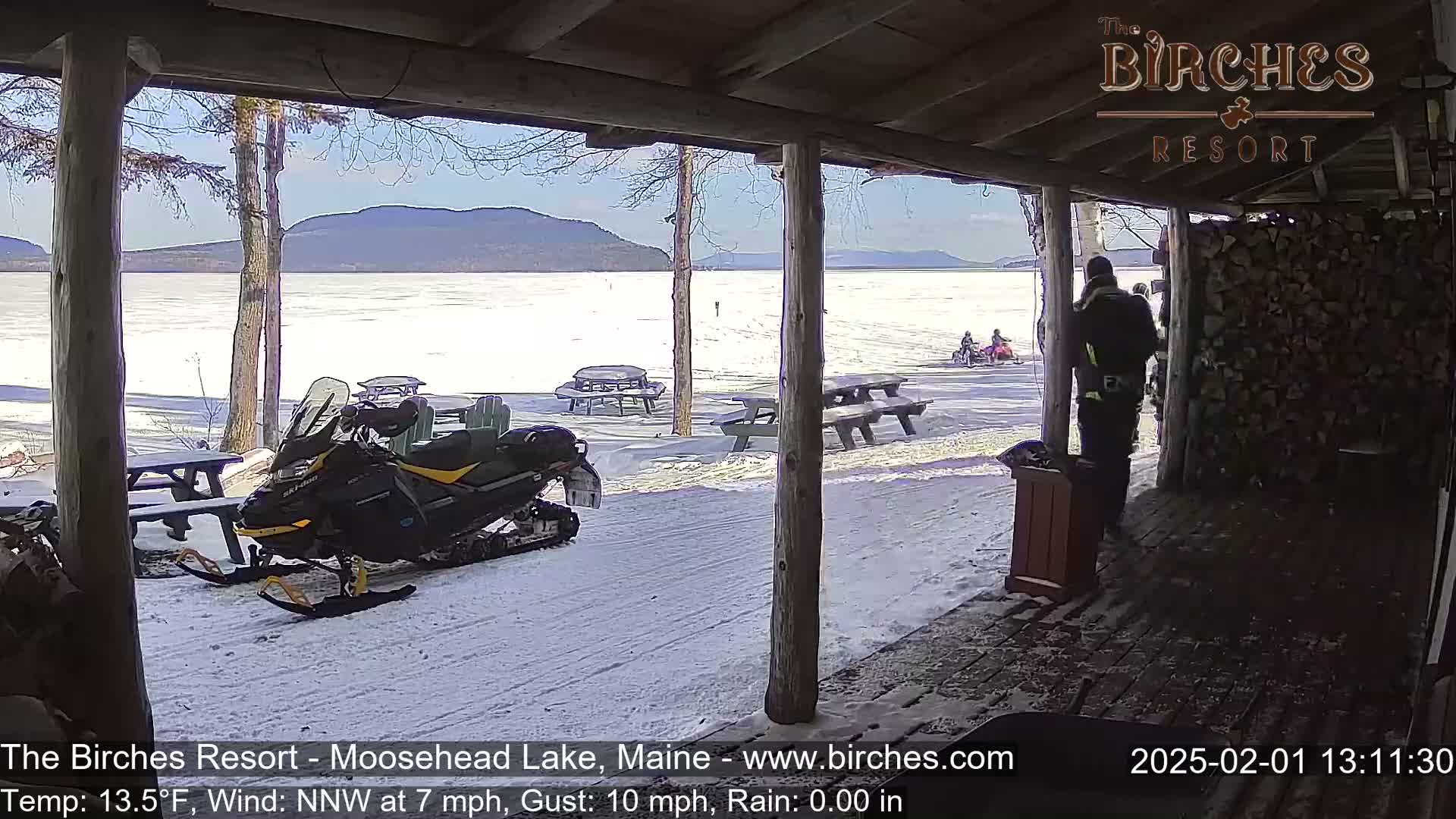 A snowmobile sits parked near picnic tables on a snow-covered lakeside, under a sunny sky with a view of distant mountains and a frozen lake.