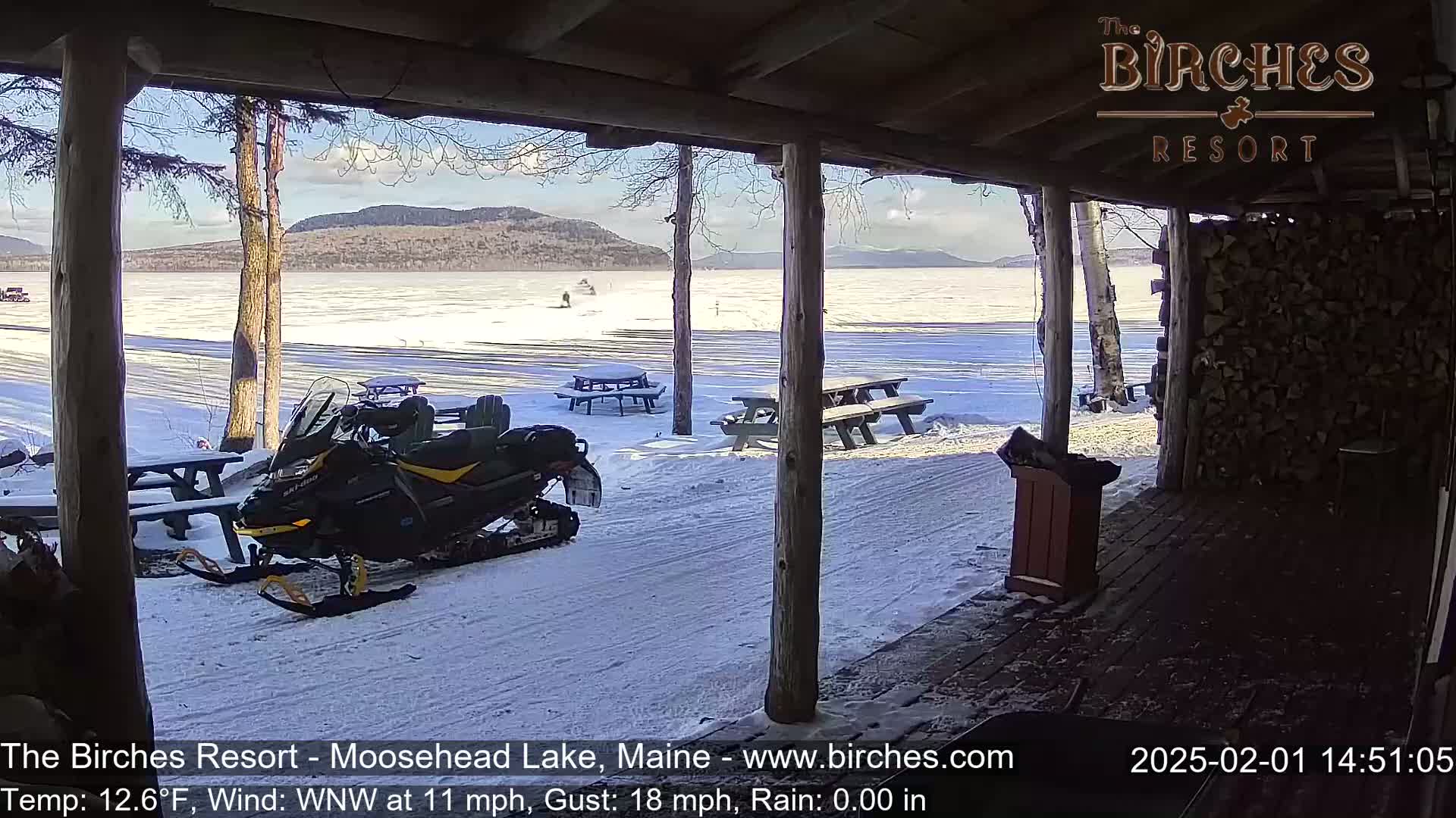 A snowmobile sits parked near picnic tables on a snow-covered lakeside area, with a mountain range visible in the background under a sunny sky.
