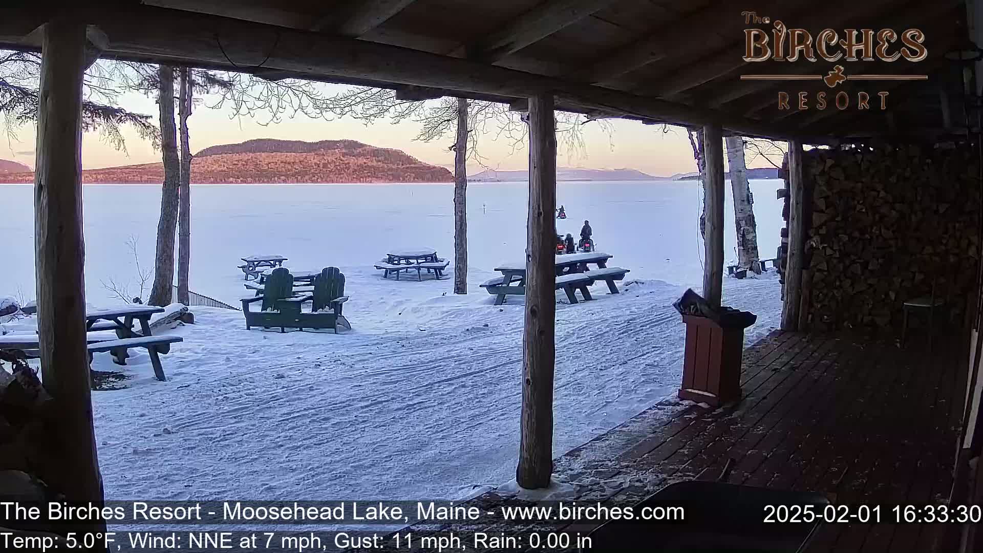 A snow-covered lakeside scene is visible from a covered porch, with picnic tables and chairs scattered across the snow, and snowmobiles on a frozen lake in the background under a twilight sky.