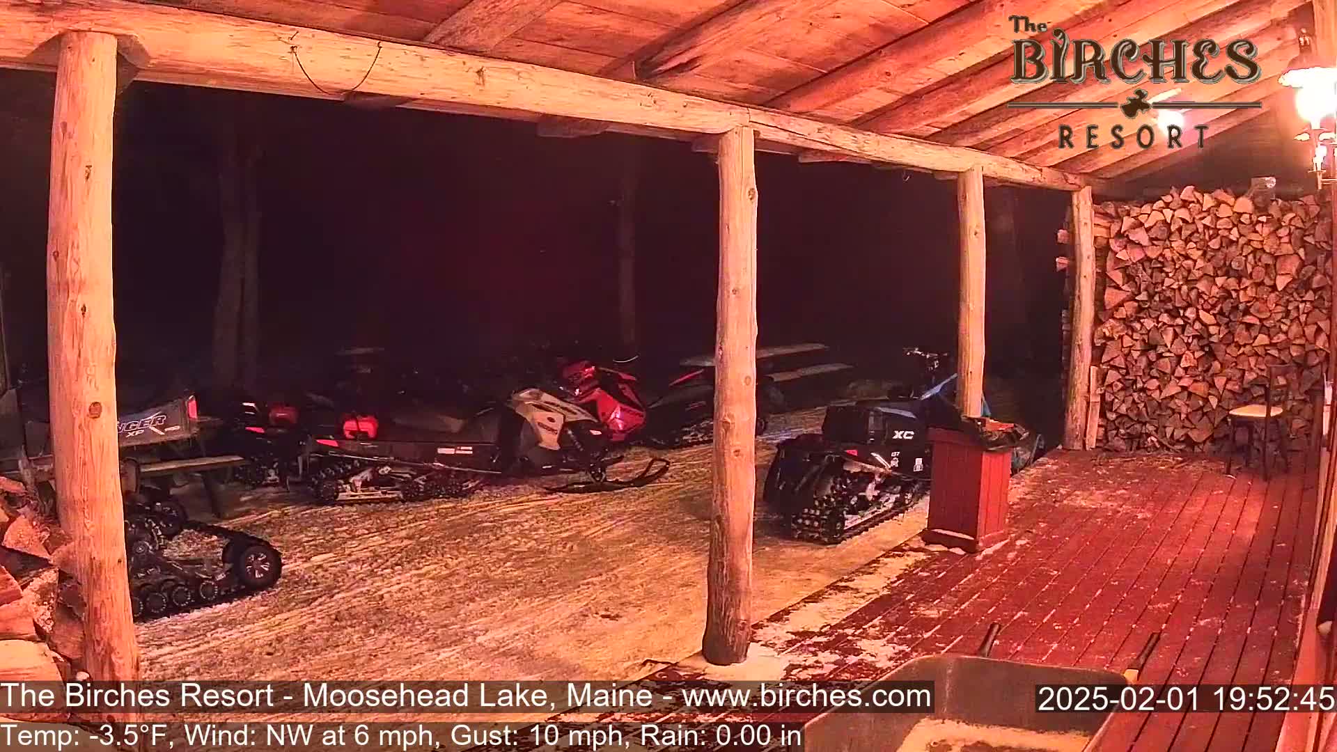 Several snowmobiles are parked under a covered porch at night, with a woodpile visible to the right and a light dusting of snow on the ground.