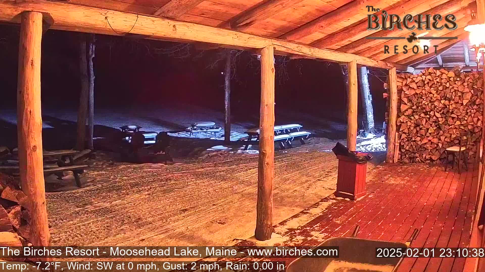 A snow-covered outdoor patio with picnic tables is visible at night from a covered porch, with a stack of firewood to the right.