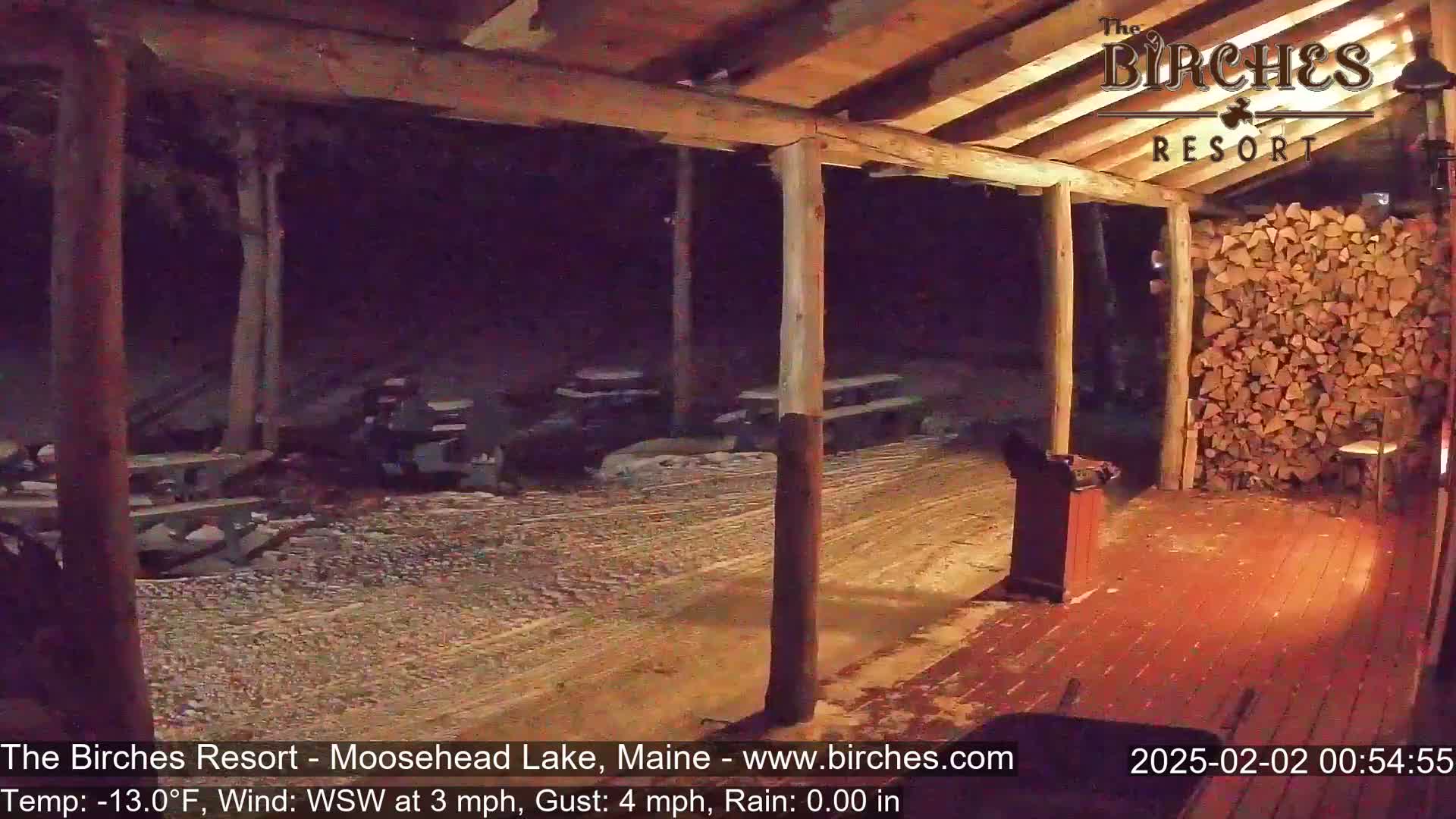 A snow-covered porch with wooden posts and a woodpile is seen at night under a covered area, with light rain.