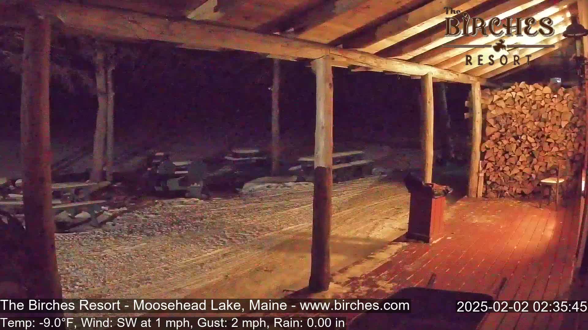 A nighttime view of a covered porch with a woodpile, picnic tables, and light snow on the ground during rainy conditions.