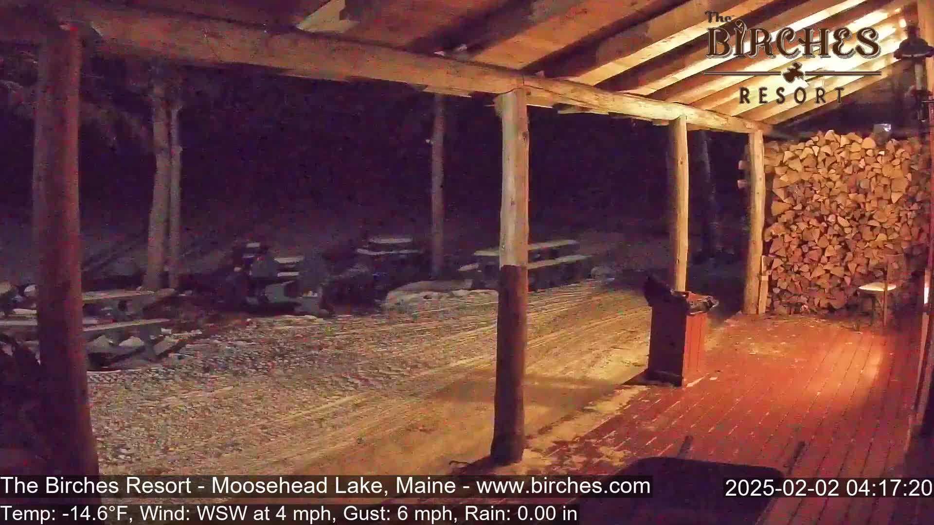 A nighttime view of a covered porch with picnic tables, a woodpile, and a lightly snow-covered ground, under a breezy, cold (-14.6°F) and rainless sky.