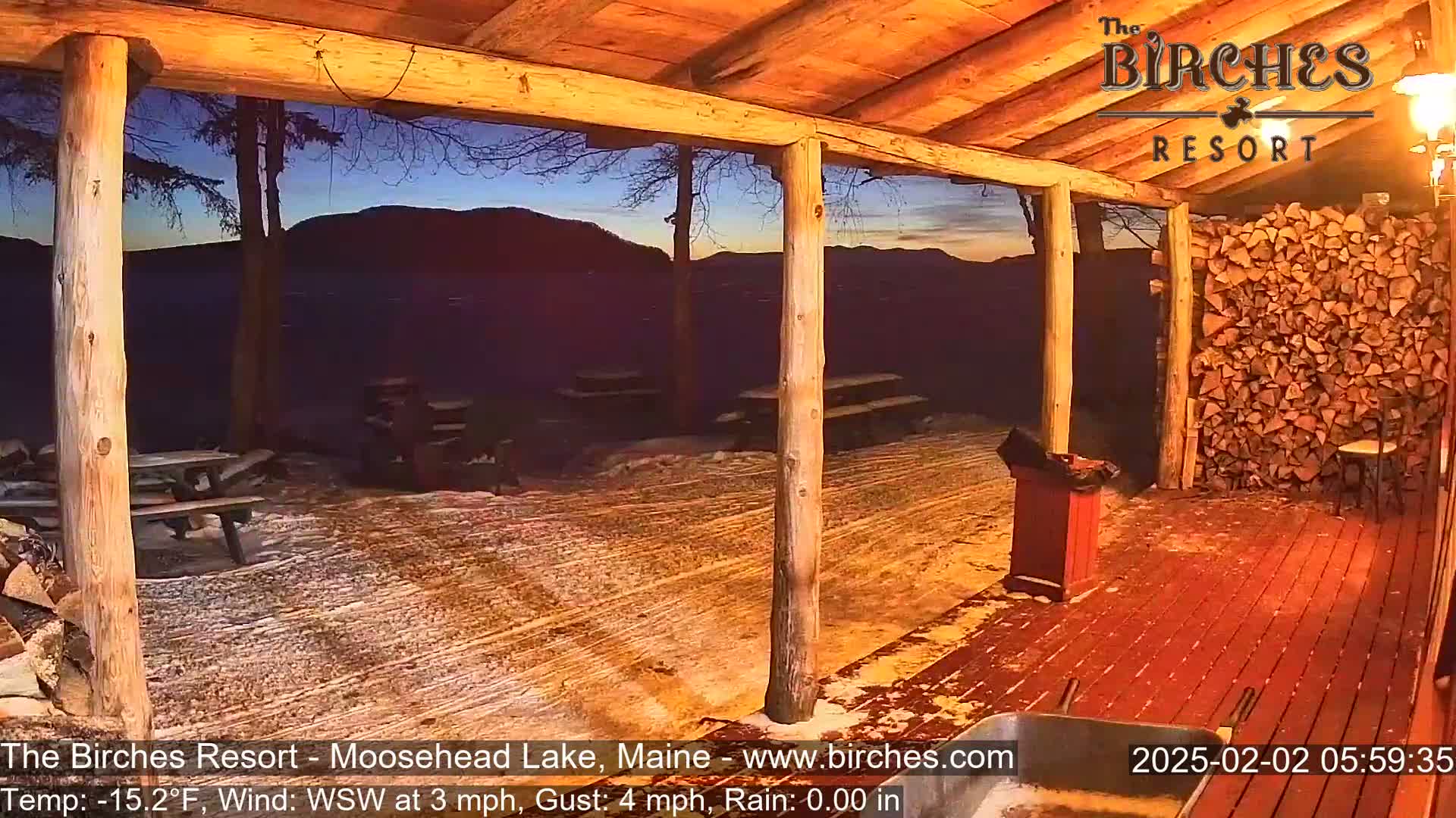 A snow-covered wooden porch with picnic tables and a woodpile overlooks a mountain range under a twilight sky during a light rain.