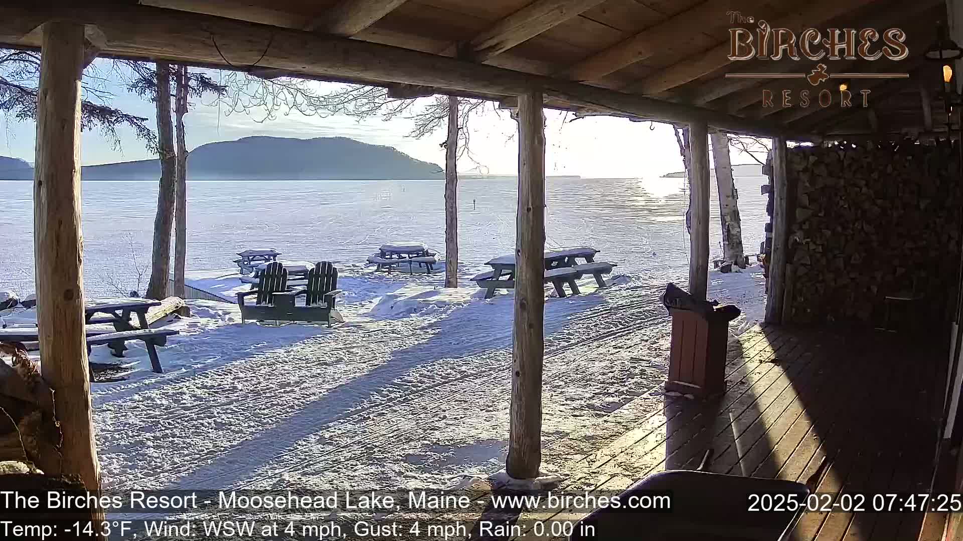 A snow-covered lakeside patio with picnic tables and Adirondack chairs is viewed from a wooden porch on a sunny, cold morning.