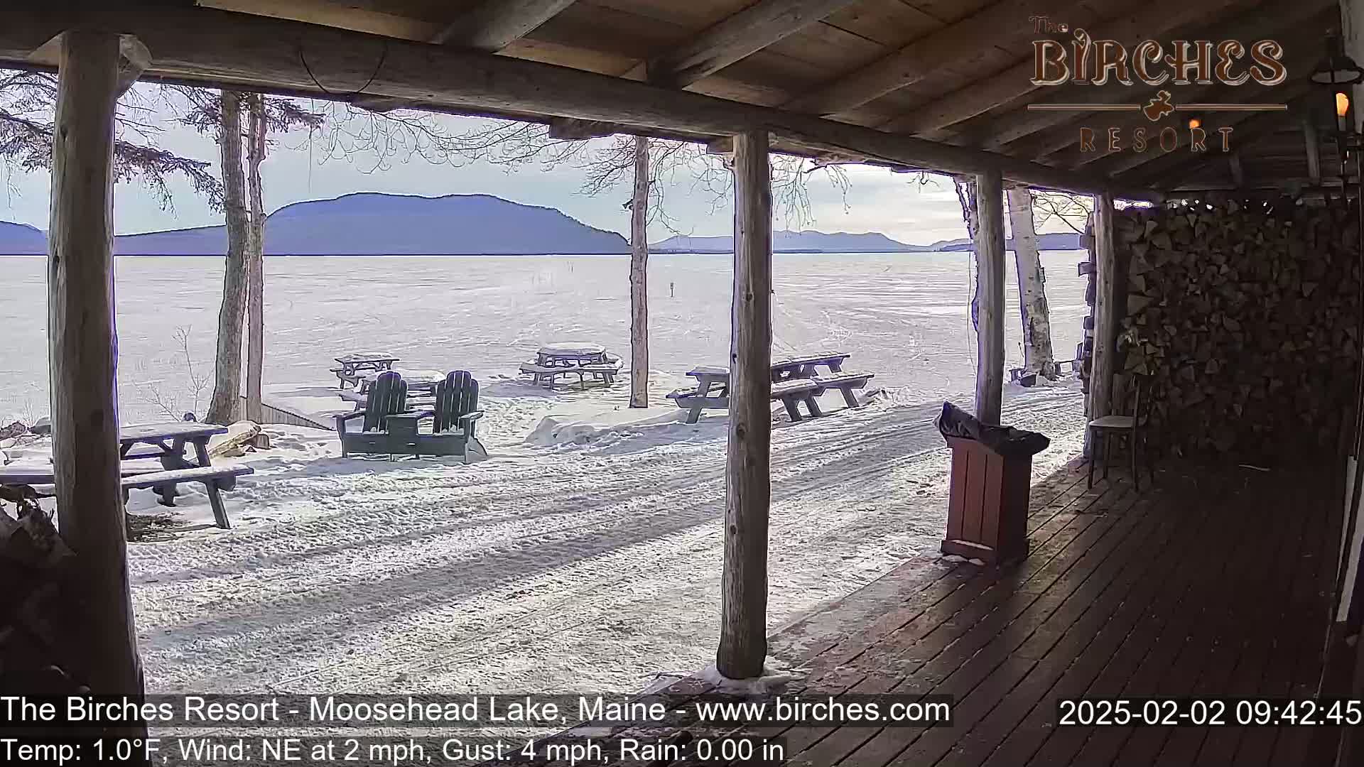 A snow-covered lakeside patio with picnic tables and Adirondack chairs is viewed from a covered porch on a partly sunny, cold day.