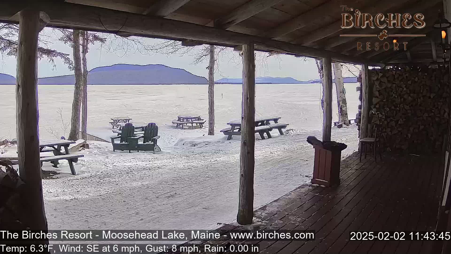 A snow-covered lakeside patio with picnic tables and Adirondack chairs offers a view of a frozen lake and distant mountains under a cloudy sky.