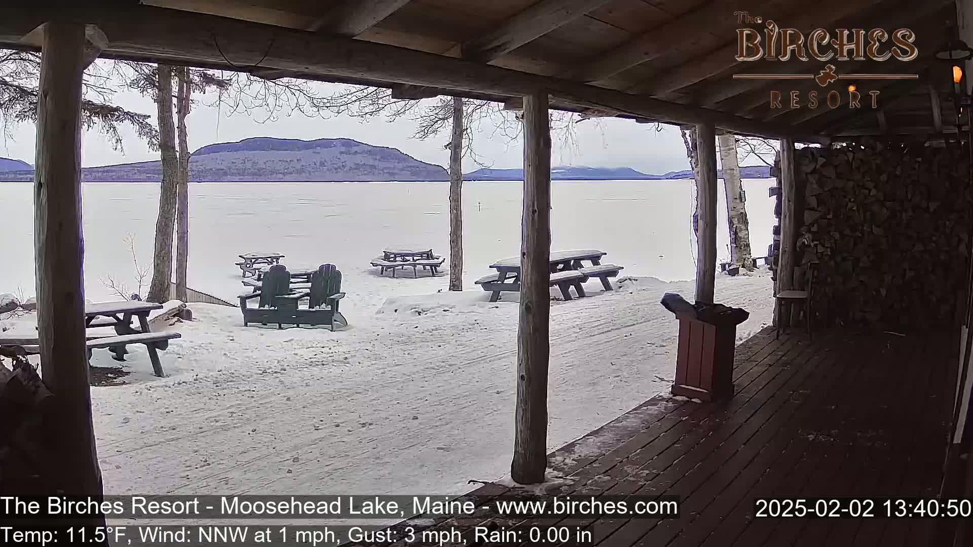 A snow-covered lakeside patio with picnic tables and Adirondack chairs is viewed from a wooden porch overlooking a frozen lake and distant mountains under a cloudy sky.