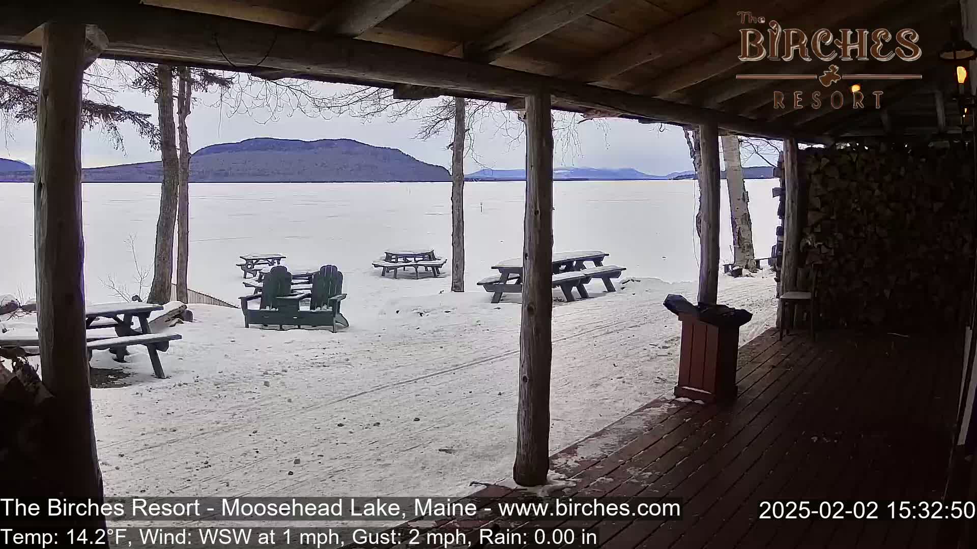 A snow-covered lakeside patio with picnic tables and Adirondack chairs is viewed from a covered porch, overlooking a frozen lake and distant mountains under a cloudy sky.