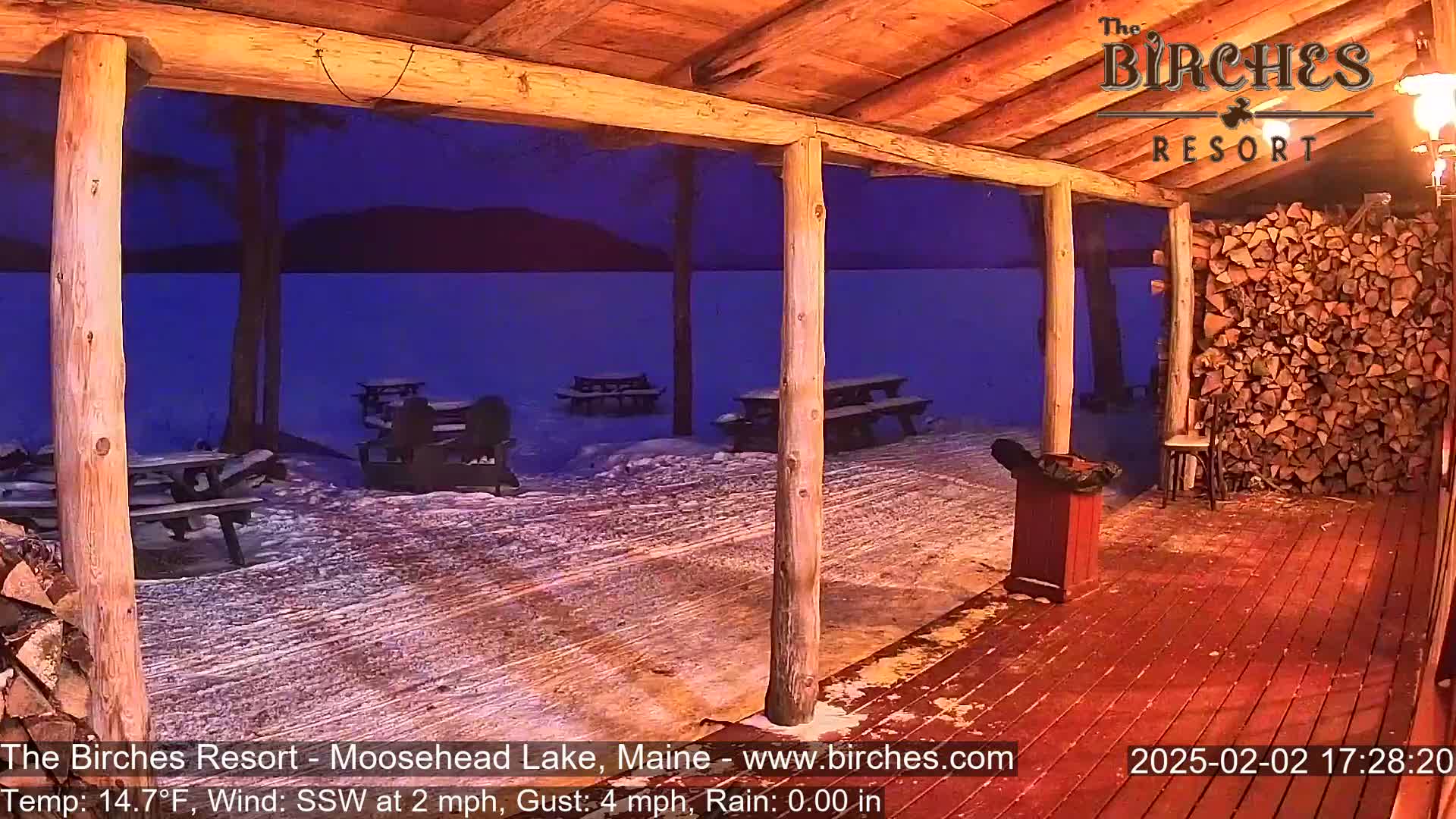 A snow-covered outdoor patio with picnic tables overlooks a dark lake under a nighttime sky with light rain.