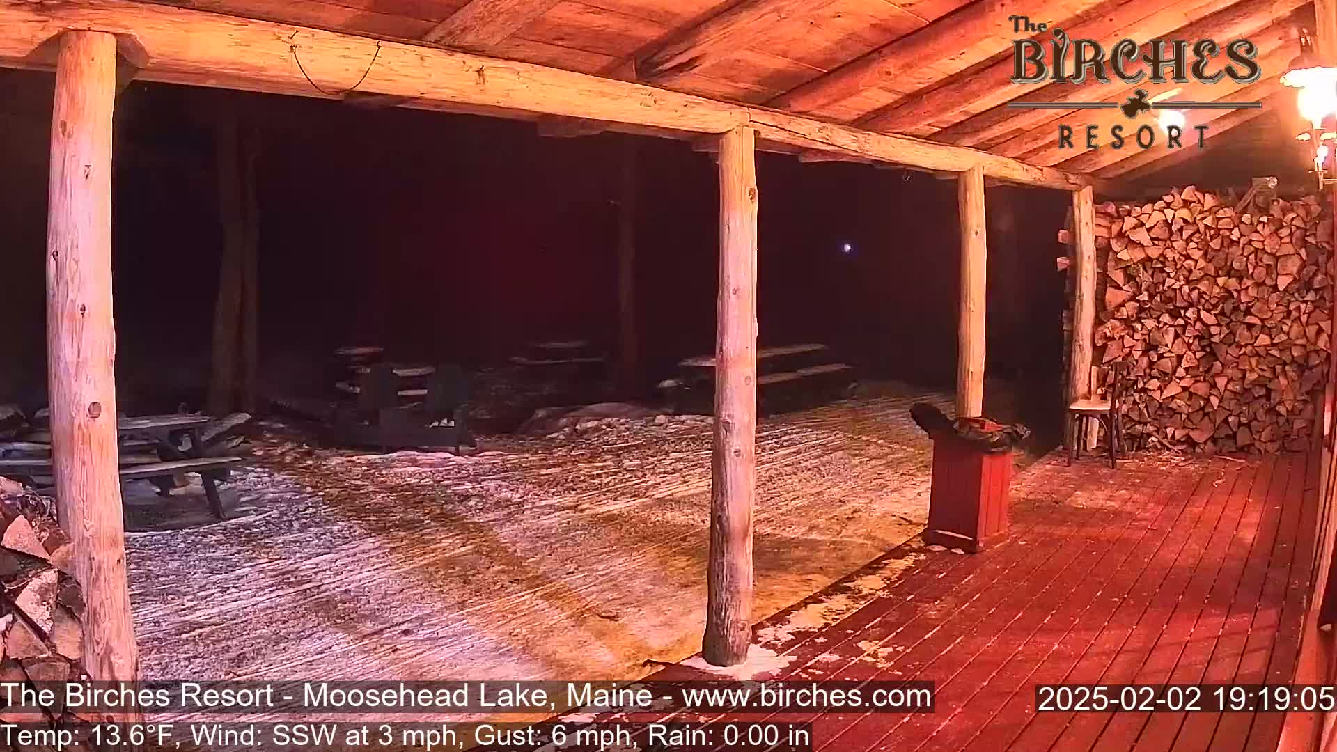 A snow-covered outdoor patio area with wooden picnic tables and a woodpile under a wooden structure at night, in rainy conditions with a south-southwesterly wind.