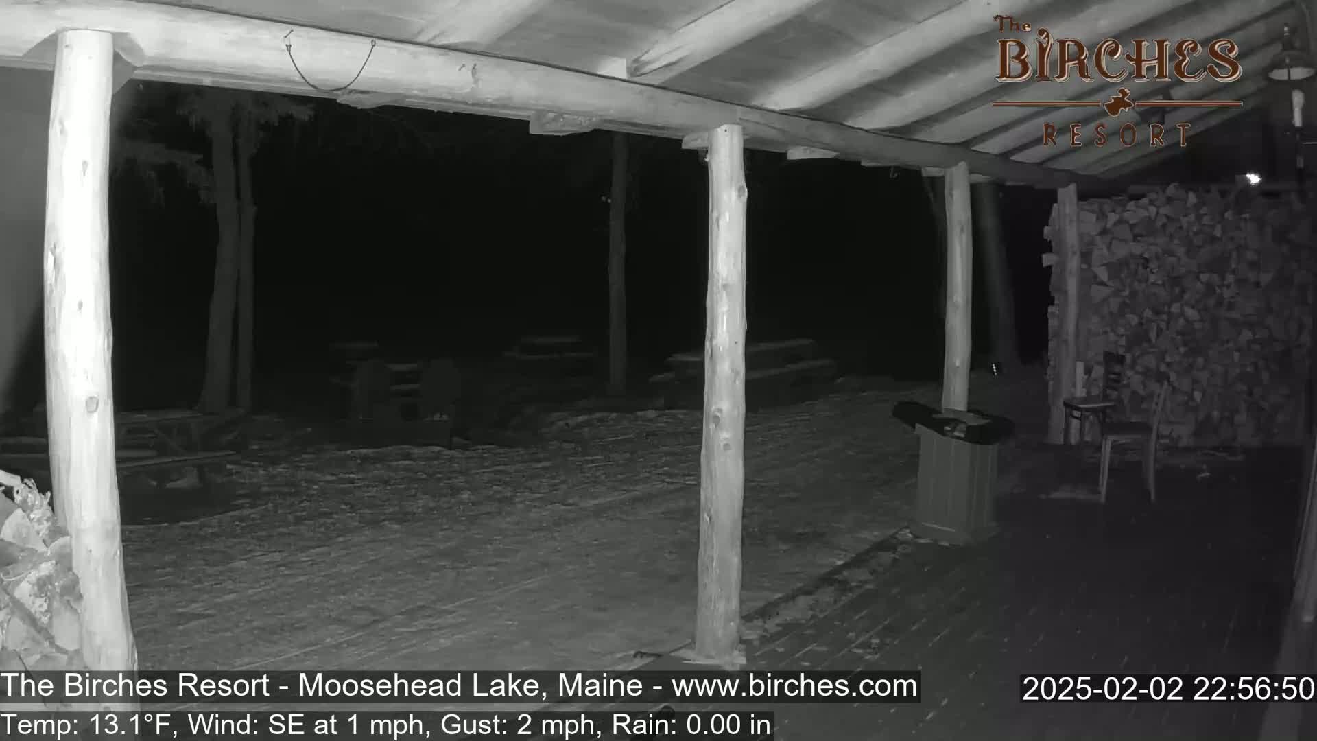 A snow-covered outdoor patio area at night, partially sheltered by a wooden structure with supporting posts, is illuminated by a low-light camera.