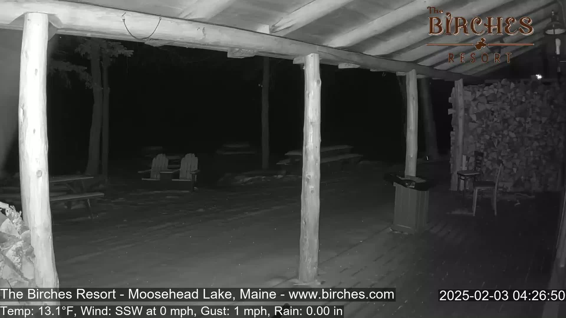 A nighttime, snow-covered outdoor patio with wooden posts, picnic tables, and chairs, under a covered porch area.