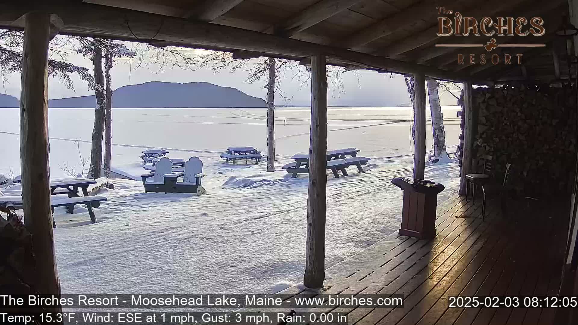 A snow-covered lakeside patio with picnic tables and chairs is seen from a covered porch, overlooking a frozen lake and distant mountains under a partly cloudy sky.