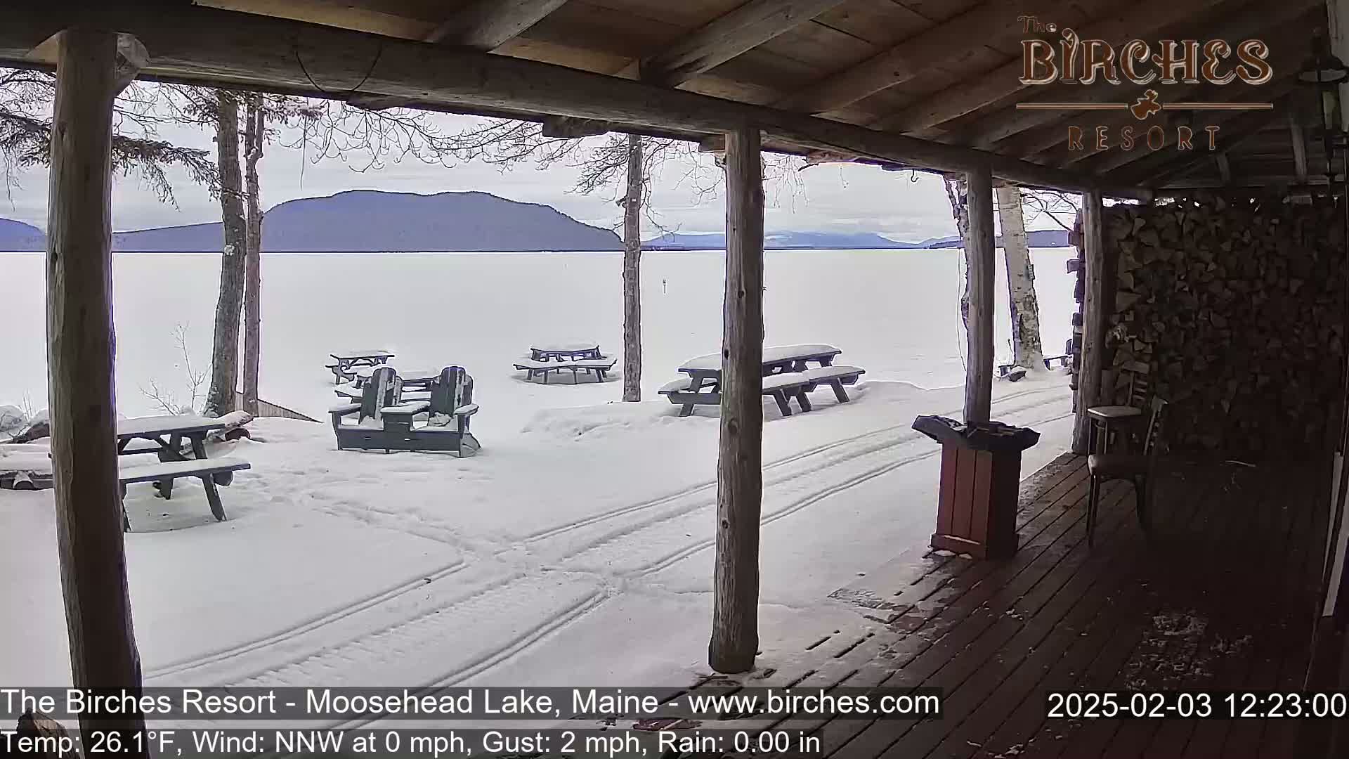 A snow-covered porch overlooks a frozen lake and distant mountains under a cloudy sky.