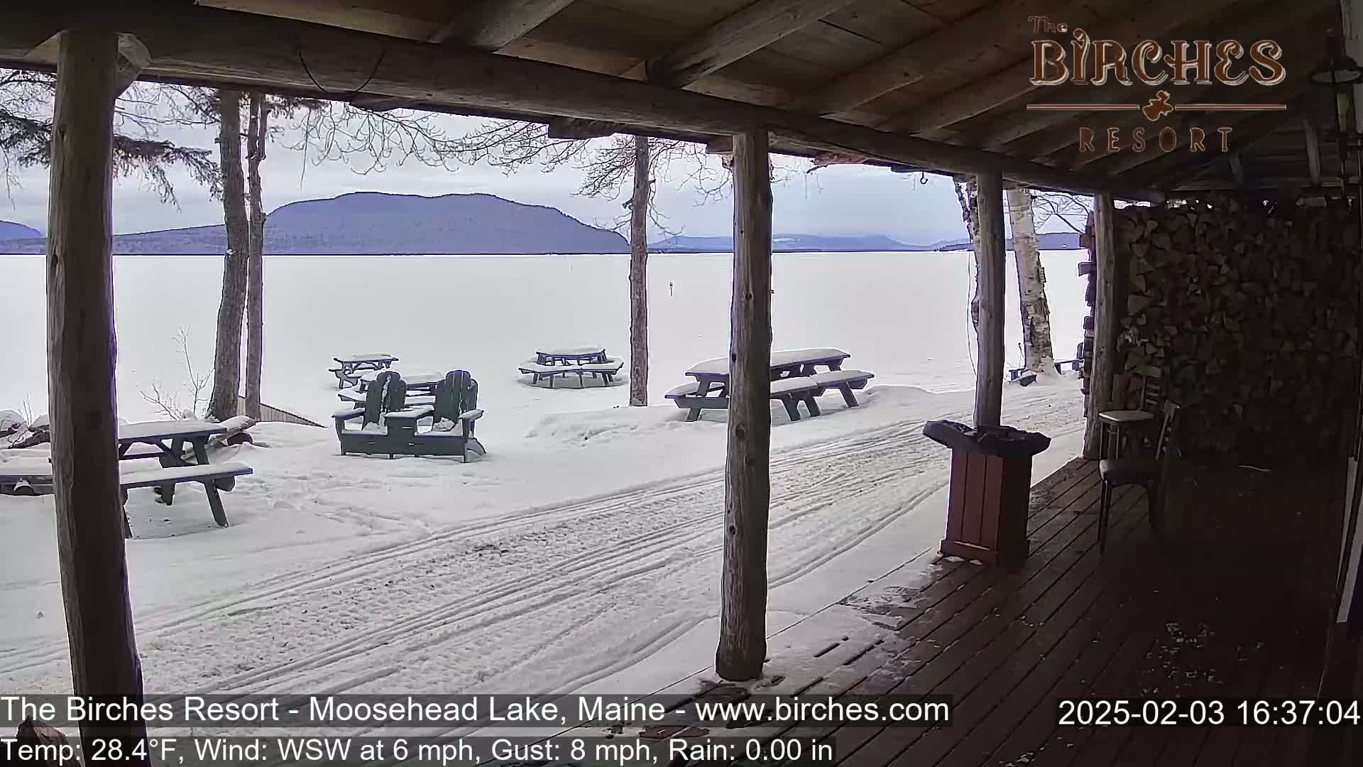 A snow-covered lakeside scene is visible from a wooden porch, with picnic tables and Adirondack chairs scattered across the snowy ground.