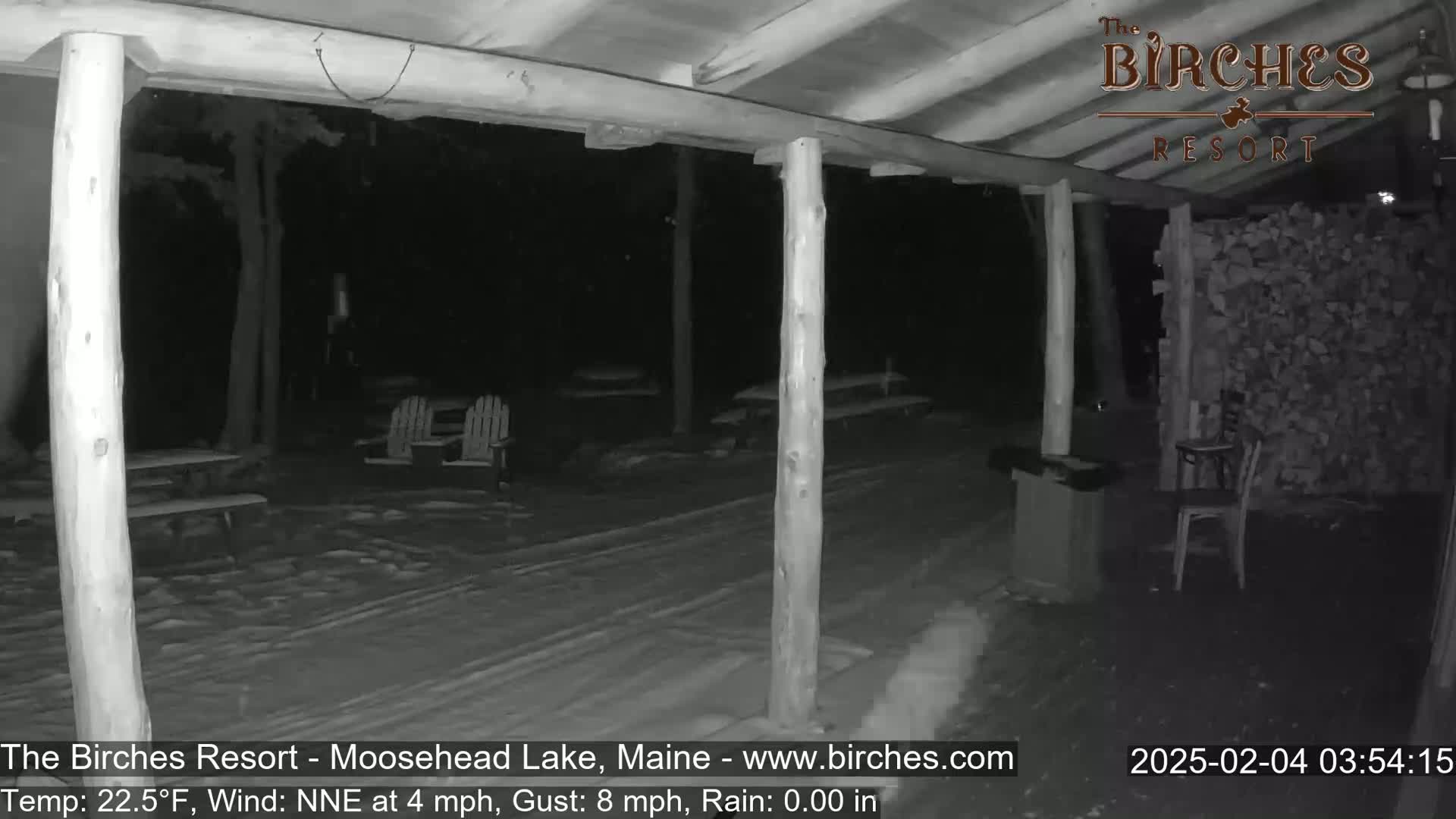 A snow-covered outdoor area at night is viewed from under a covered porch, with picnic tables and chairs visible in the distance under a slightly rainy sky.