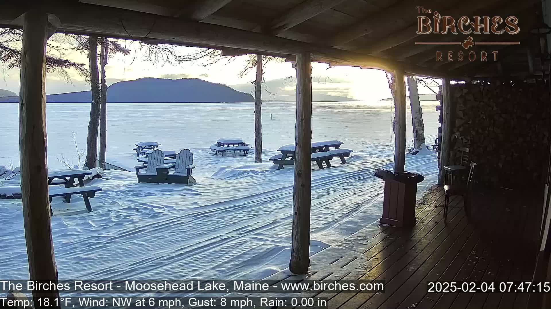 A snow-covered lakeside patio with picnic tables and Adirondack chairs is viewed from a wooden porch during a sunrise, with a light northwest wind and minimal rain.