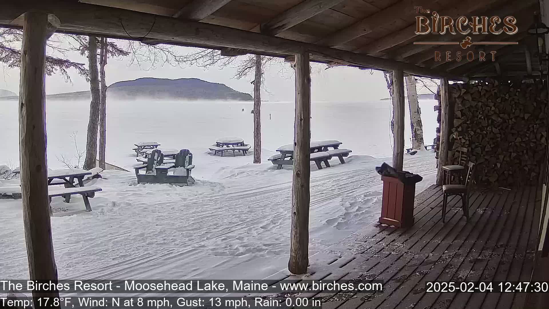 A snow-covered outdoor patio with picnic tables and chairs overlooks a foggy lake and distant mountains in windy conditions.