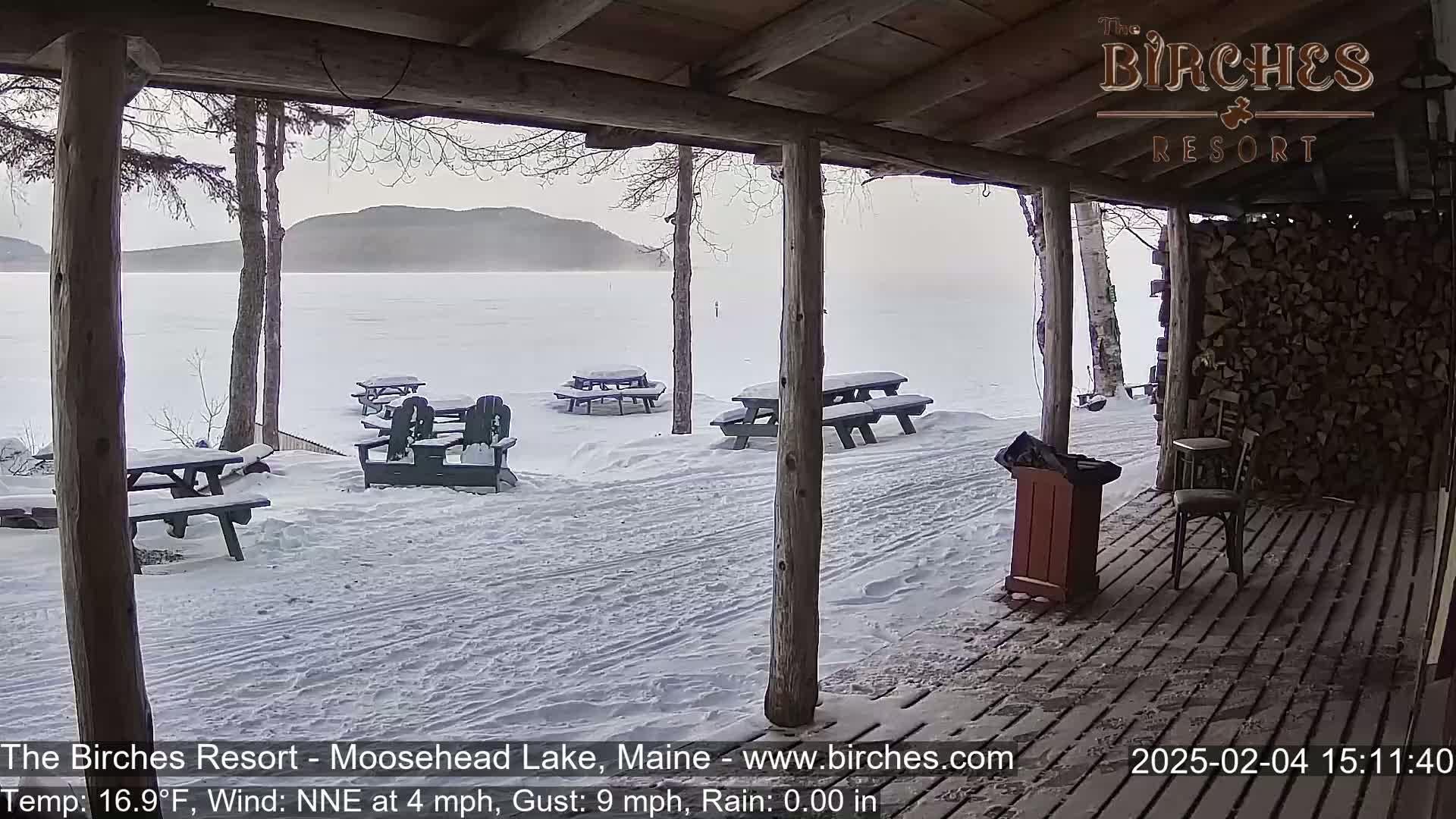 A snow-covered lakeside view with picnic tables and chairs is seen from a wooden porch in rainy, windy weather.