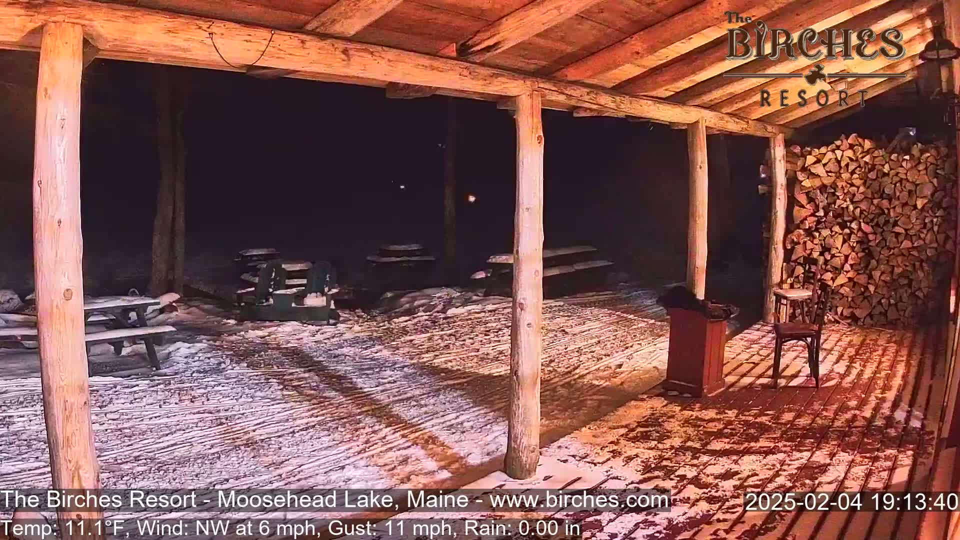 A snow-covered outdoor patio area with picnic tables is partially sheltered under a wooden structure at night.