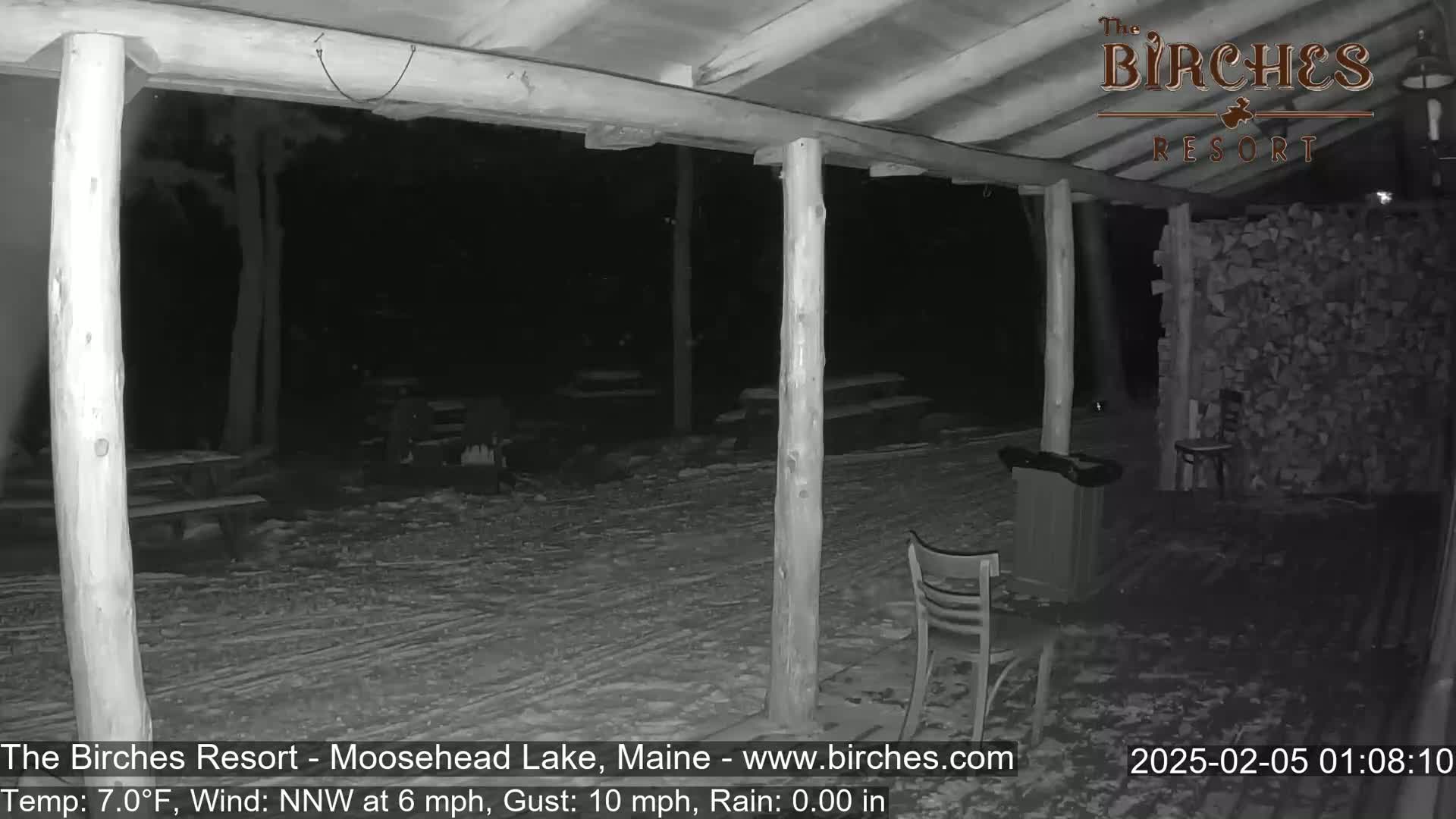 A snow-covered outdoor patio area at night, partially sheltered by a wooden structure, with a few chairs and a pile of firewood visible.