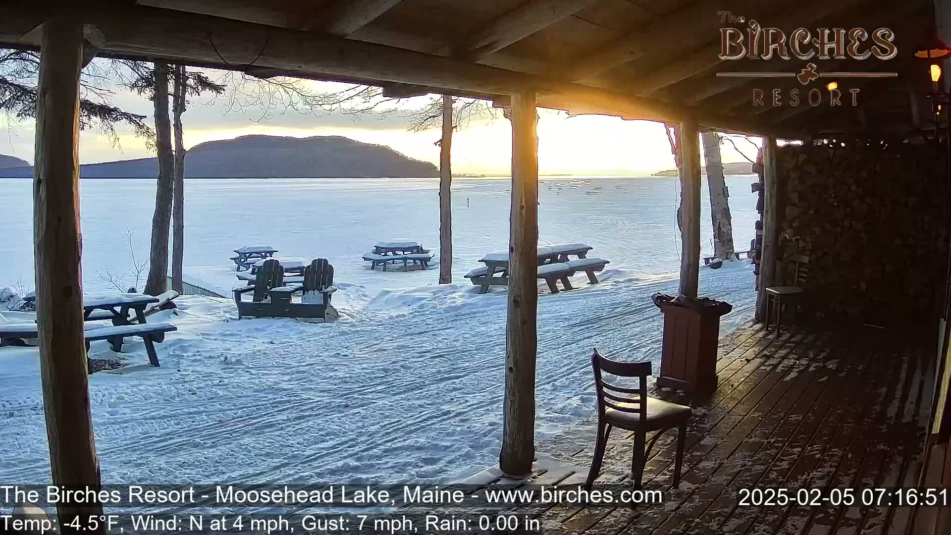 A snowy lakeside view is seen from a covered porch, with picnic tables and chairs scattered across the snow-covered ground, and a partly cloudy sunset over a frozen lake.