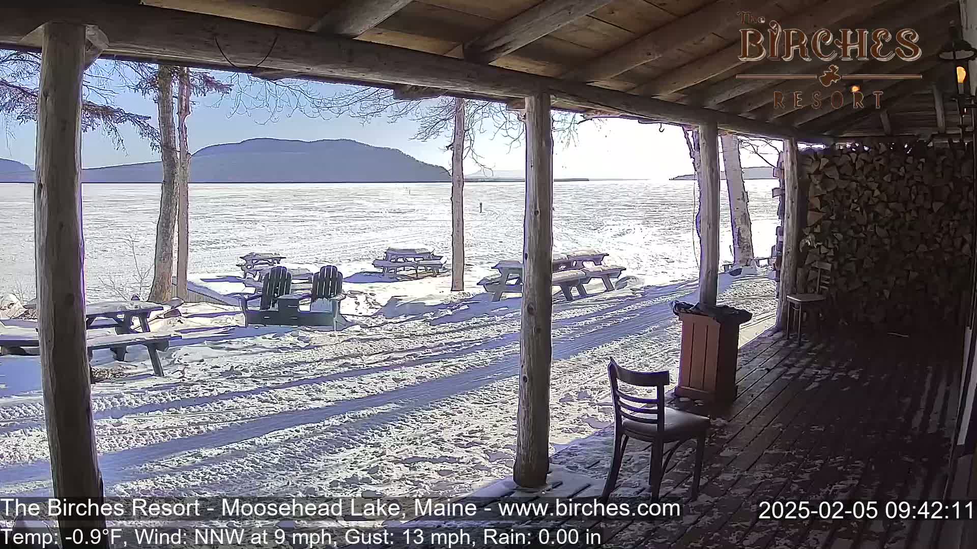 A snow-covered lakeside patio with picnic tables and chairs is viewed from a wooden porch on a sunny, windy day.