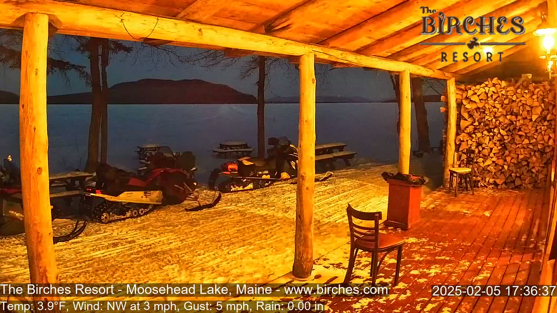 Snow-covered ground outside a wooden porch overlooks a calm lake under a dark night sky, with snowmobiles parked nearby.